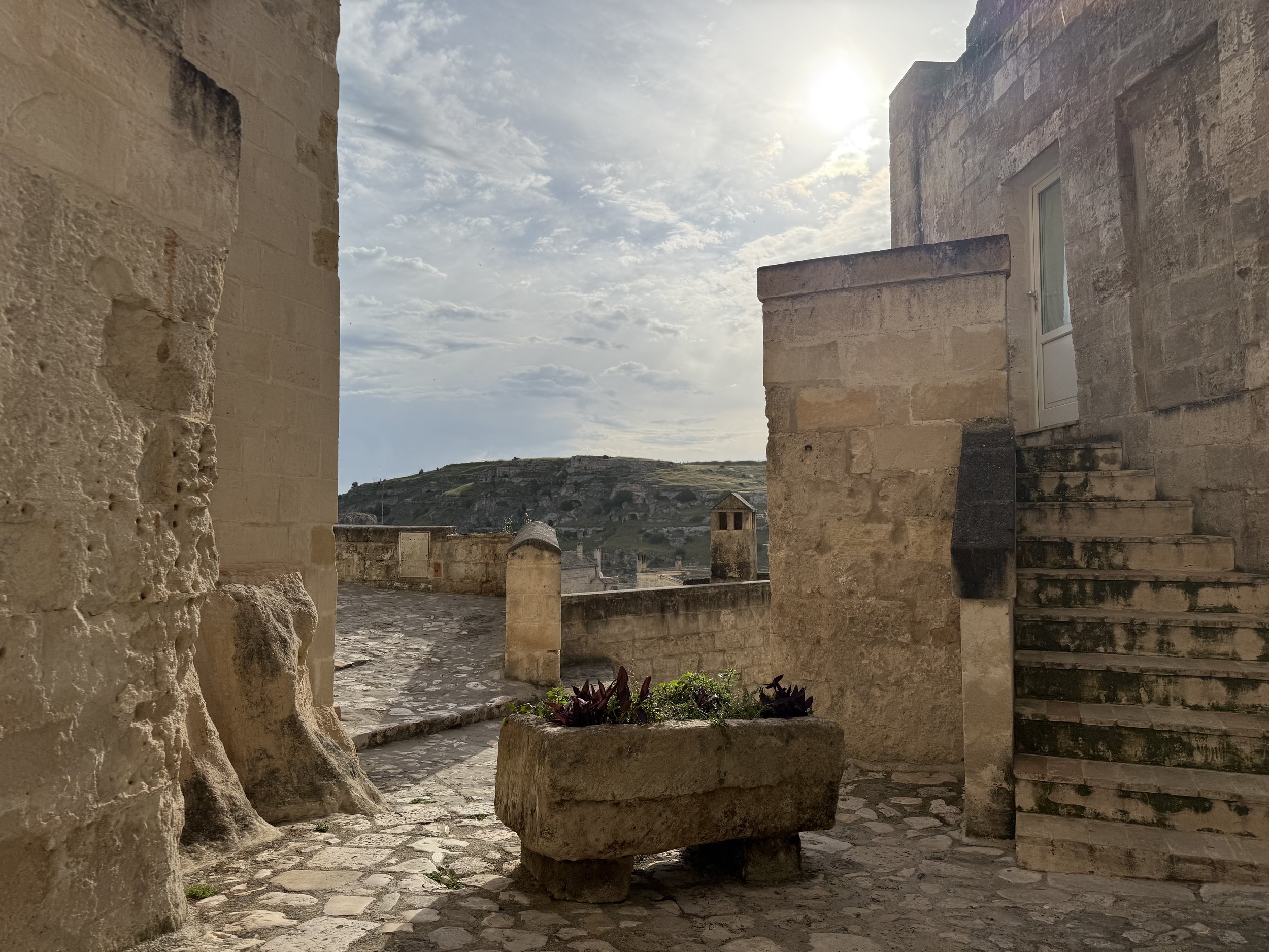 The photo shows an old stone building and historic ruins, a stone street, and a view into the distance with rolling hills and small buildings. The sky is partly cloudy with the sun shining through.