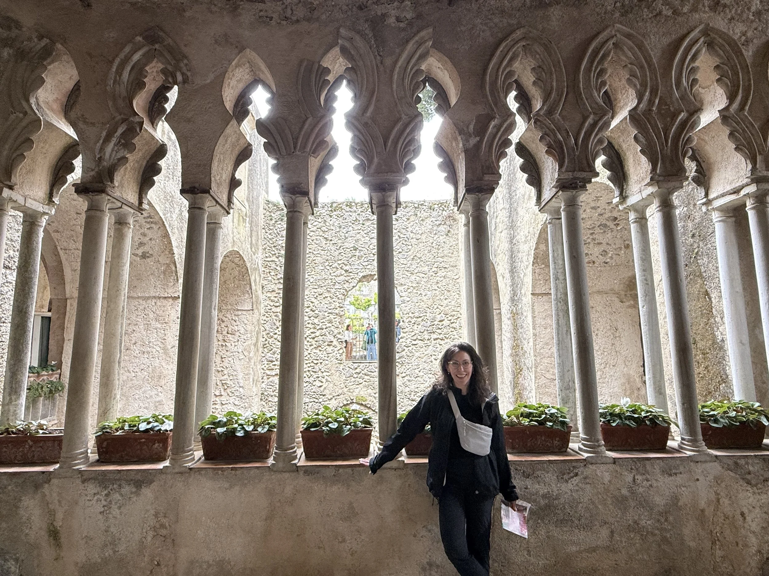 A woman with glasses and long dark hair, wearing a black jacket and white shirt, standing in front of an arched stone window with decorative columns. Several potted plants are placed on the window ledge, and sunlight streams through the openings.