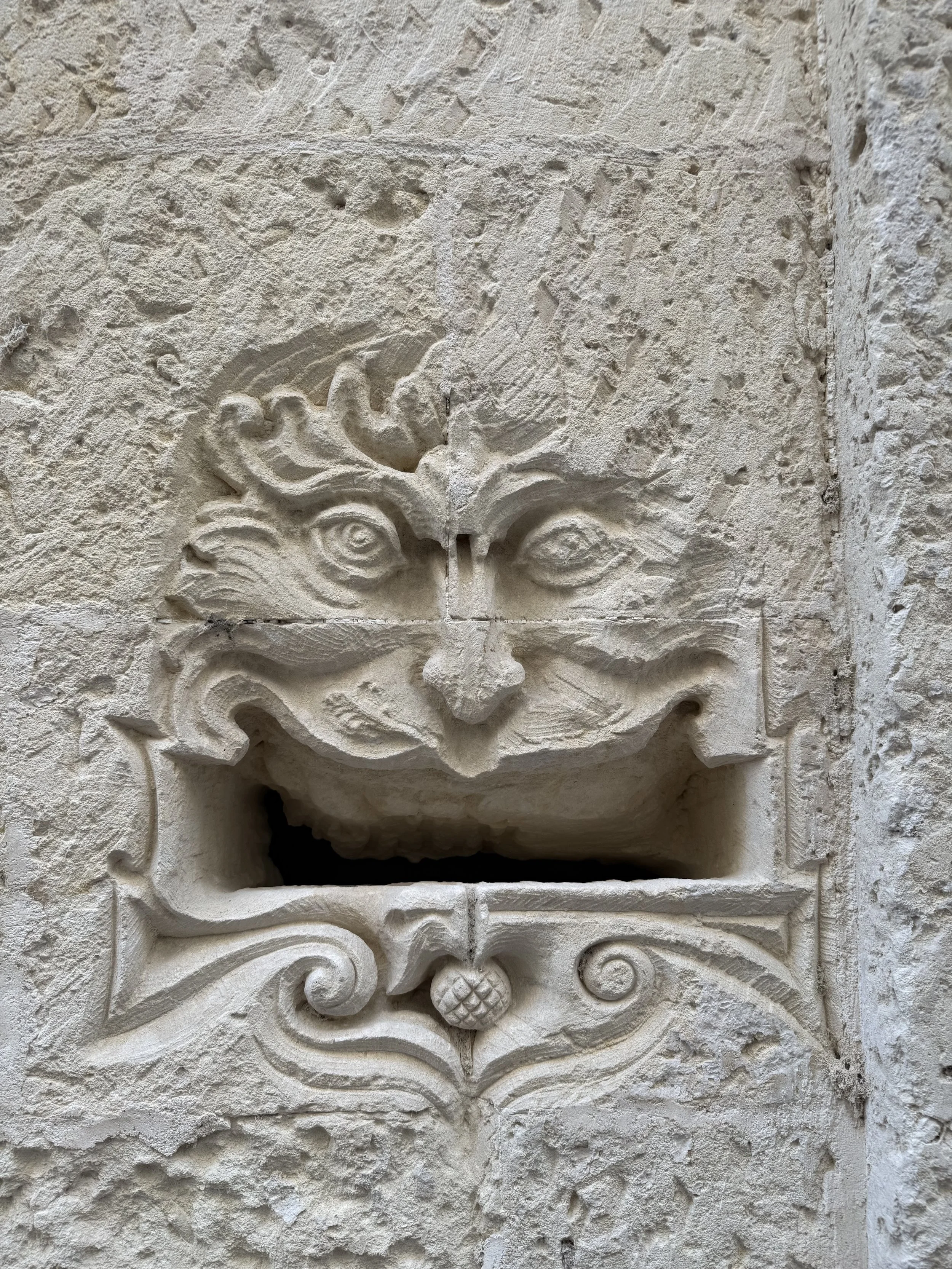 Close-up of a carved stone face relief on a textured stone wall, featuring intricate details and swirling decorative elements.
