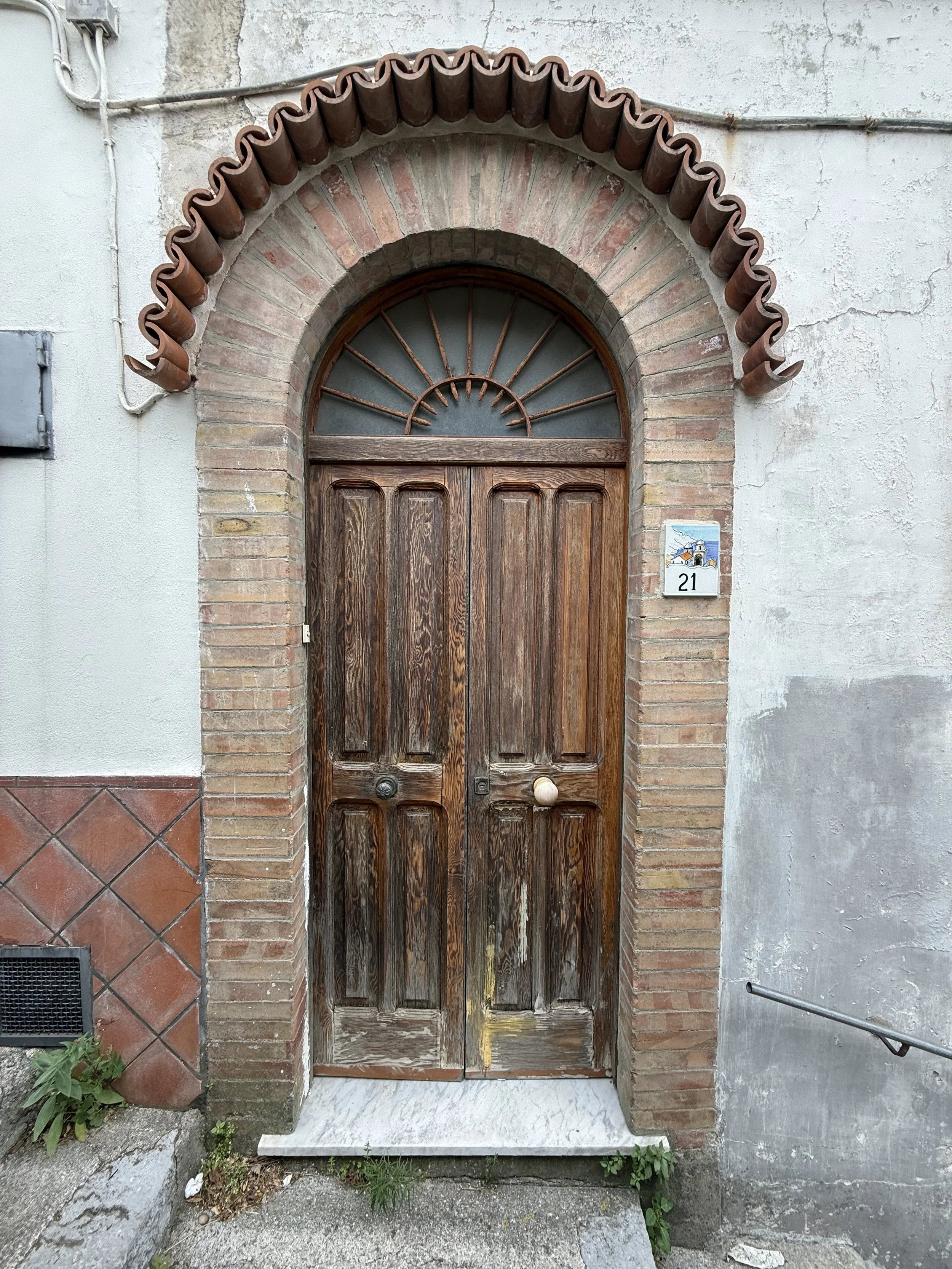 Old wooden double door with a semi-circular metal grille window above, framed by brick archway, with house number 21 on a small tiled plaque to the right.