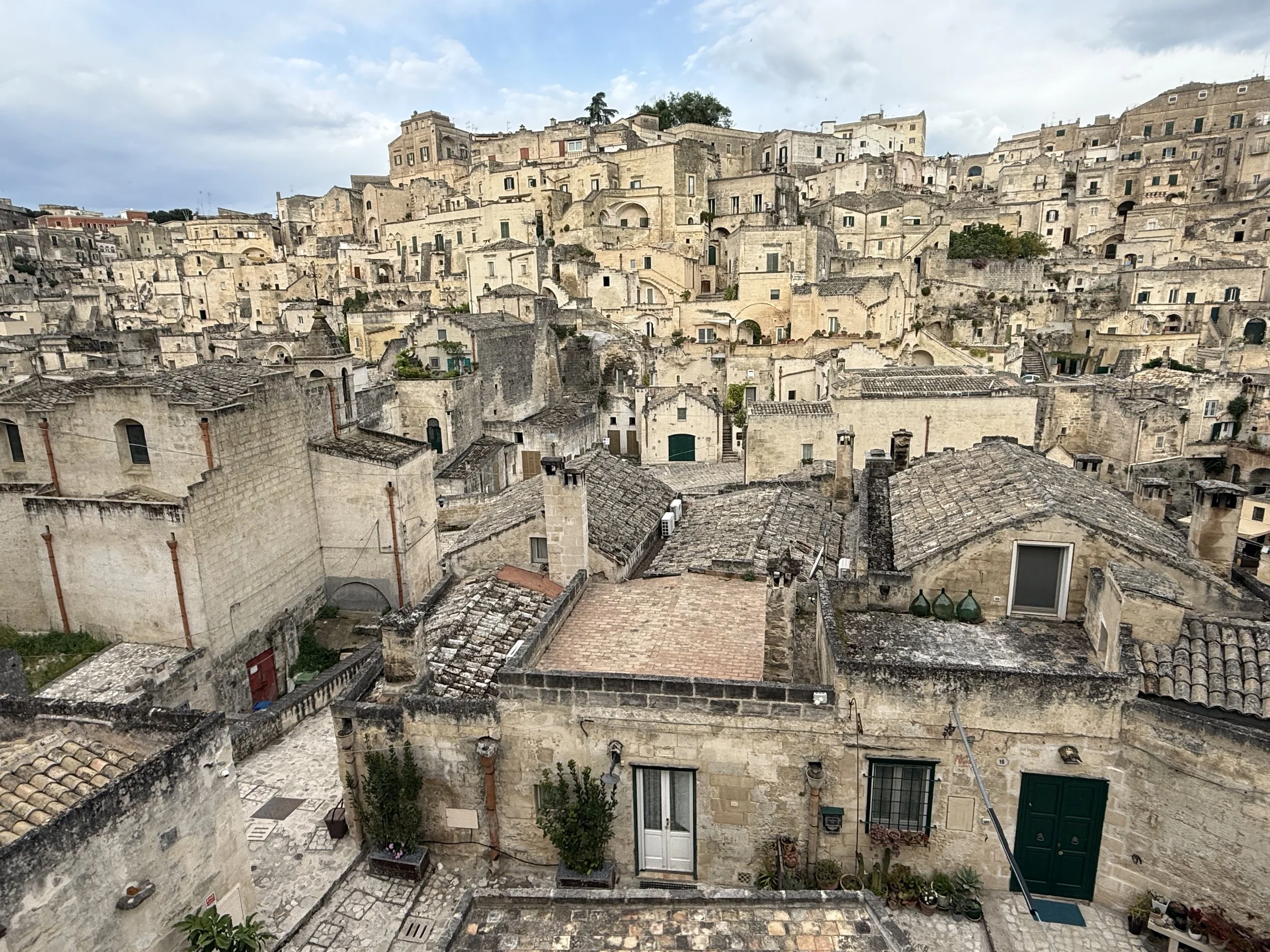 A scenic view of an ancient hillside town with densely packed stone buildings and narrow streets, with some greenery at the top and scattered trees amid the buildings.