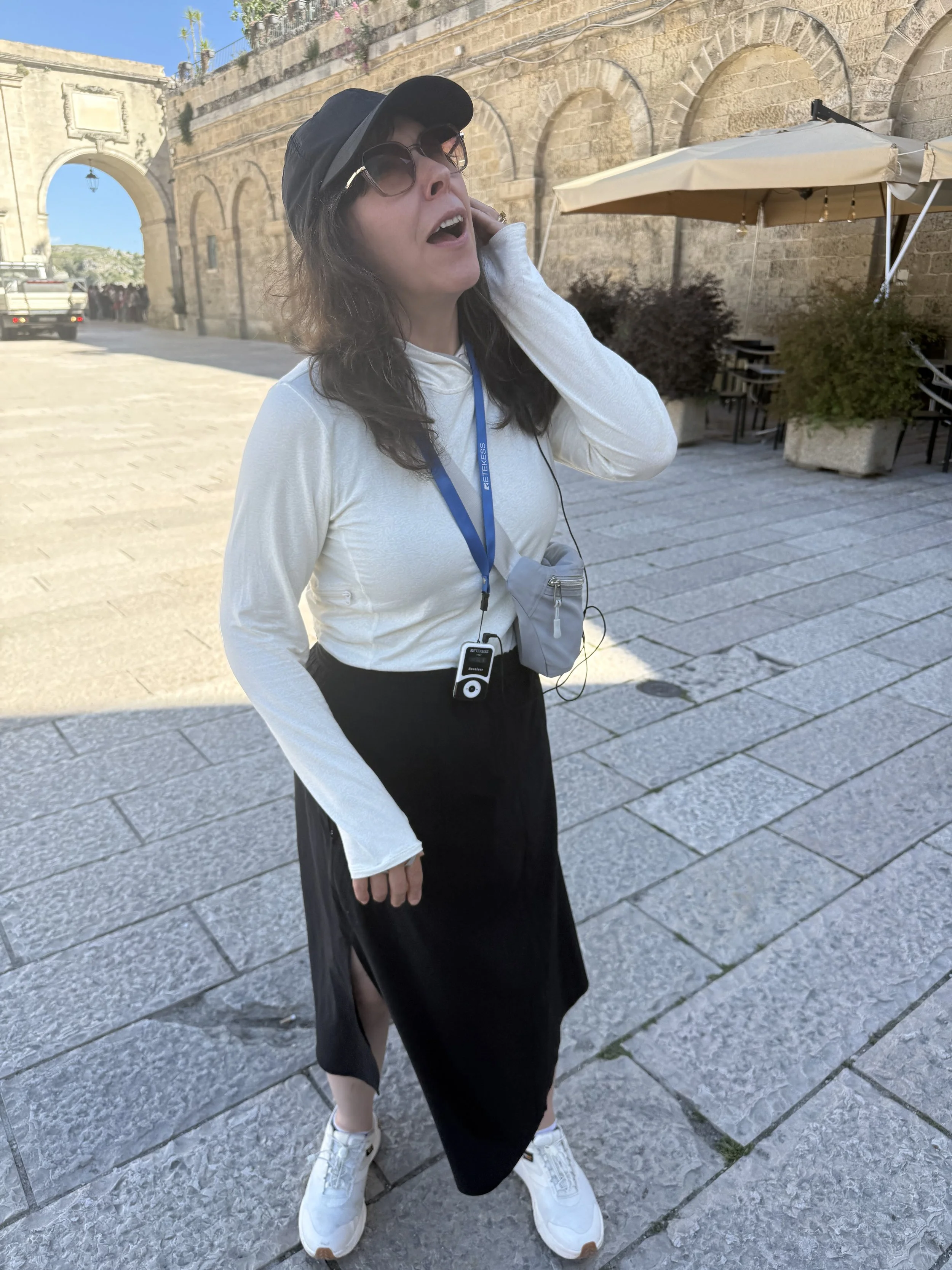 A woman with curly brown hair wearing sunglasses, a black hat, a white long-sleeve shirt, a black skirt, white sneakers, and a blue lanyard stands outdoors on a stone-paved street with an ancient stone arch and building, under a clear blue sky.
