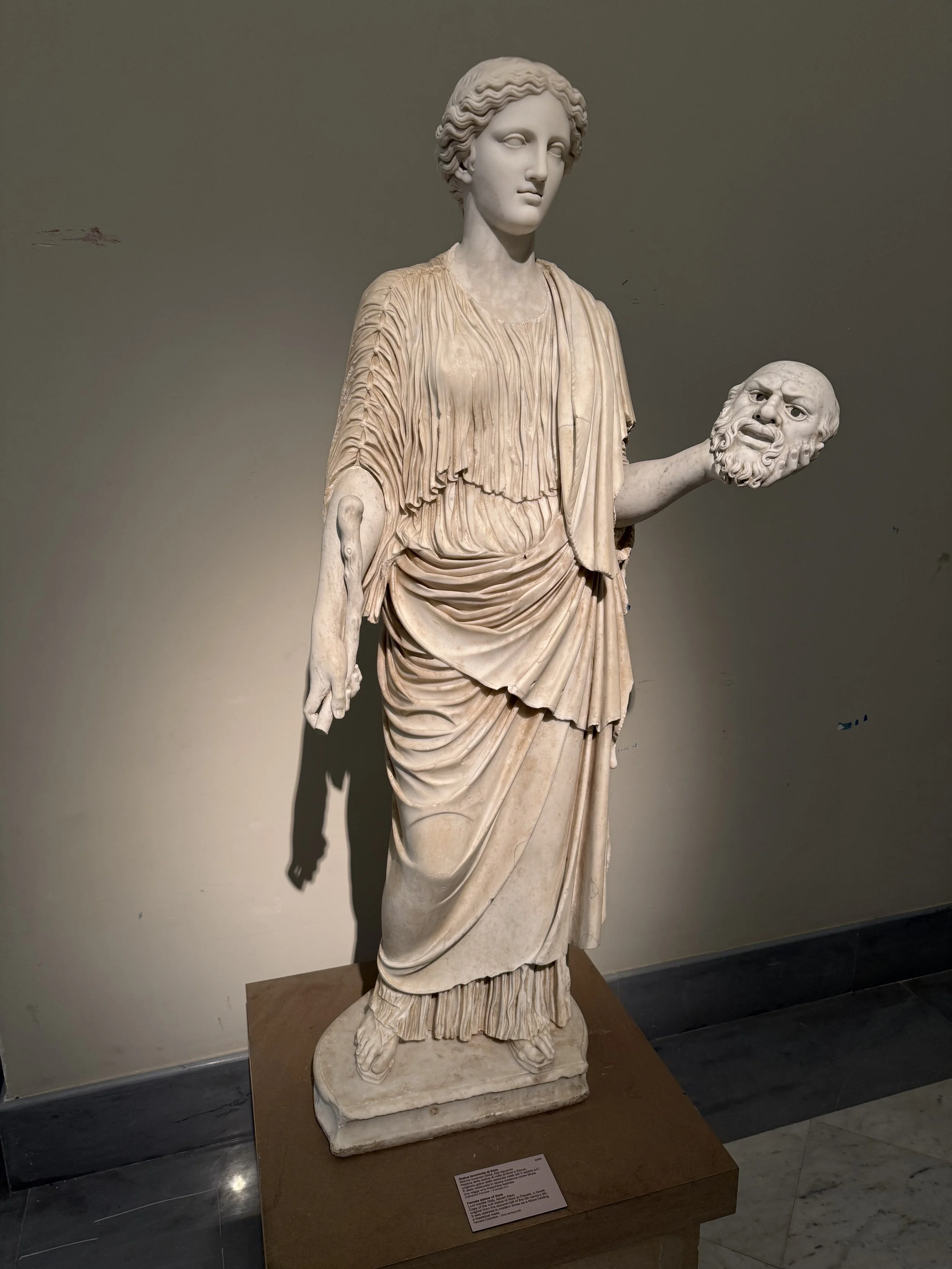Ancient marble sculpture of a standing woman holding a decapitated head in her left hand, dressed in classical draped clothing, located in a museum setting.