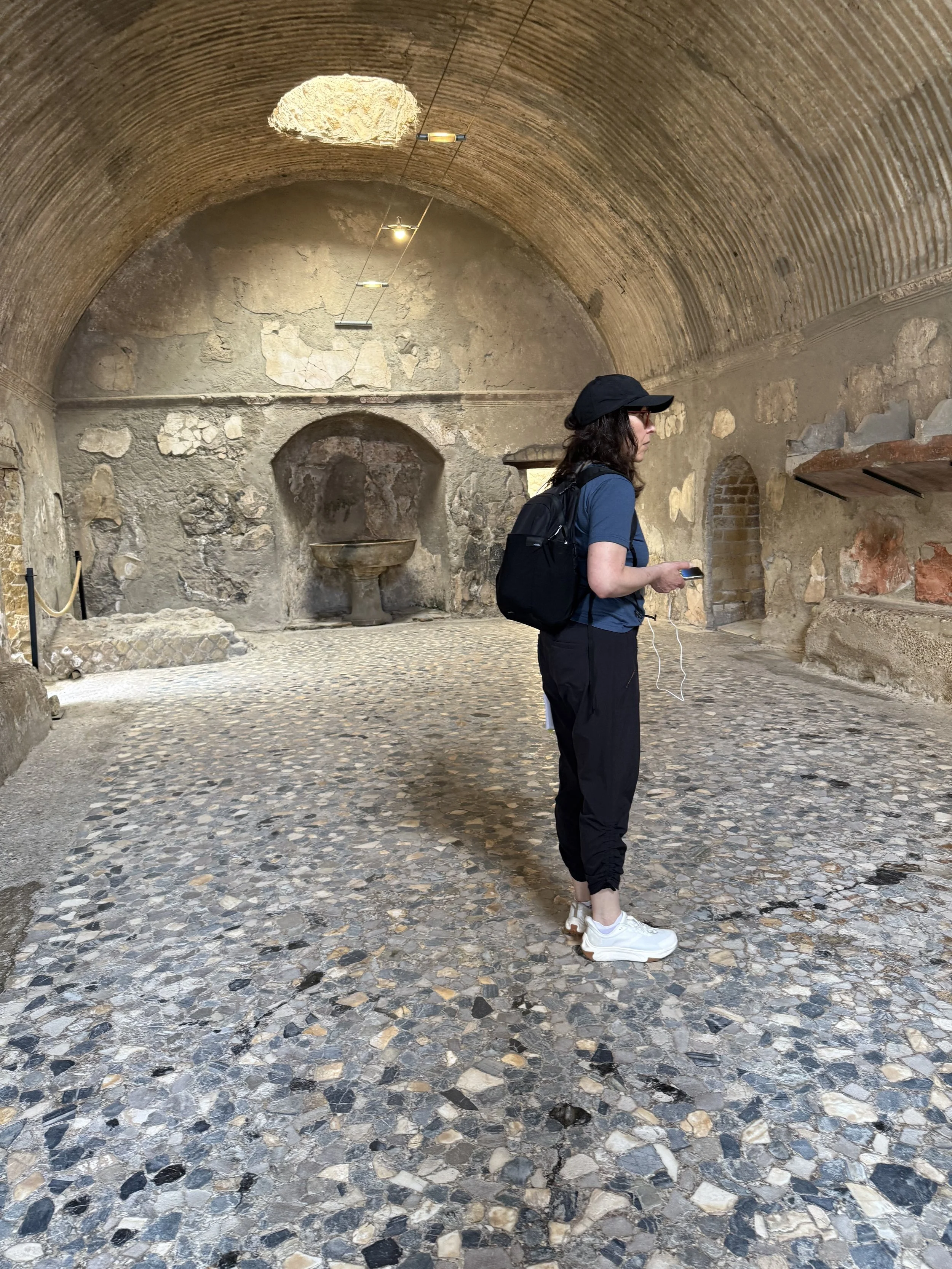 Woman standing inside ancient underground structure with stone walls and vaulted ceiling, wearing a black cap, blue shirt, black pants, white sneakers, holding a phone, and wearing a backpack.