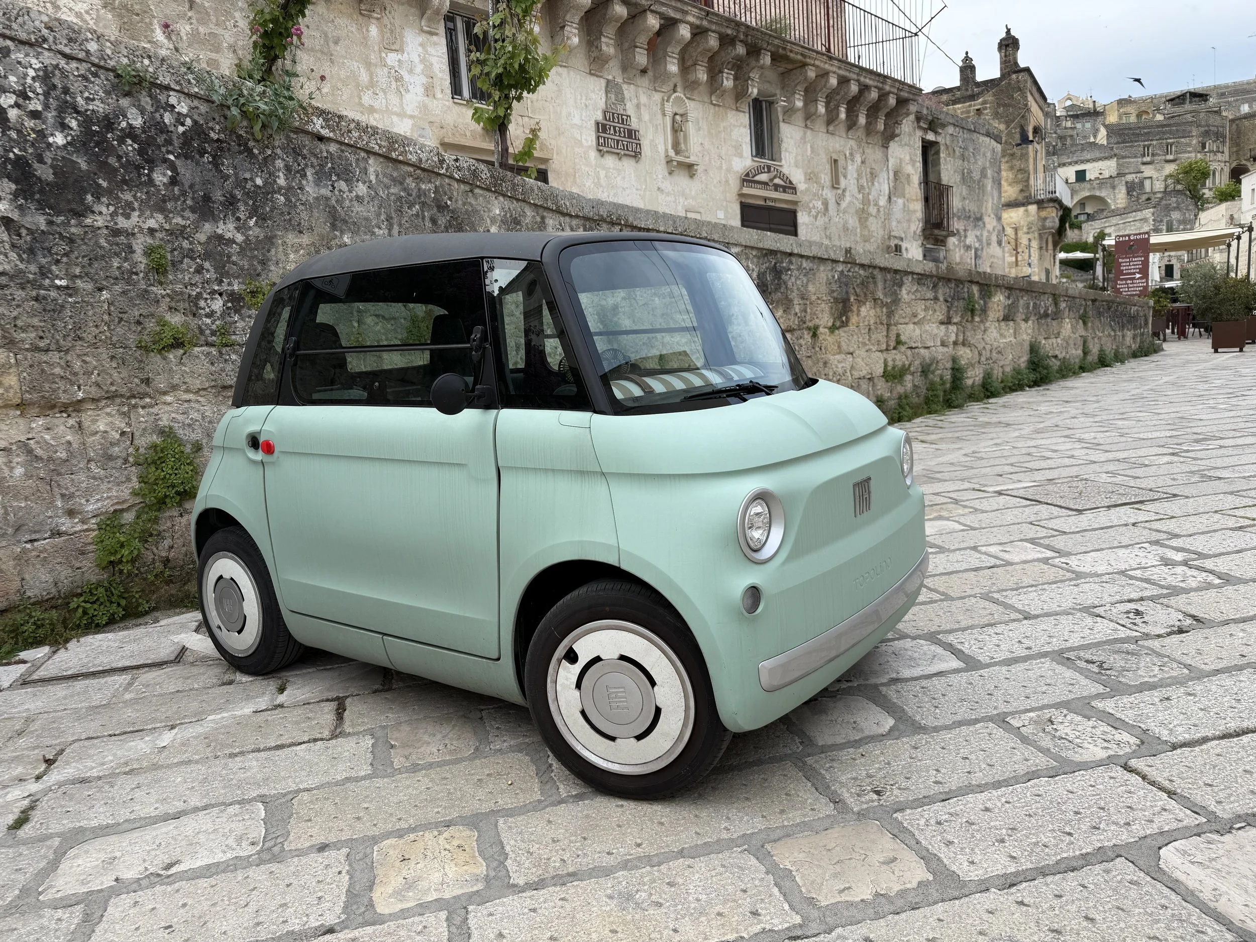 A small, light green electric vehicle with a black roof parked on a cobblestone street beside an old stone wall and historic buildings in a European town.