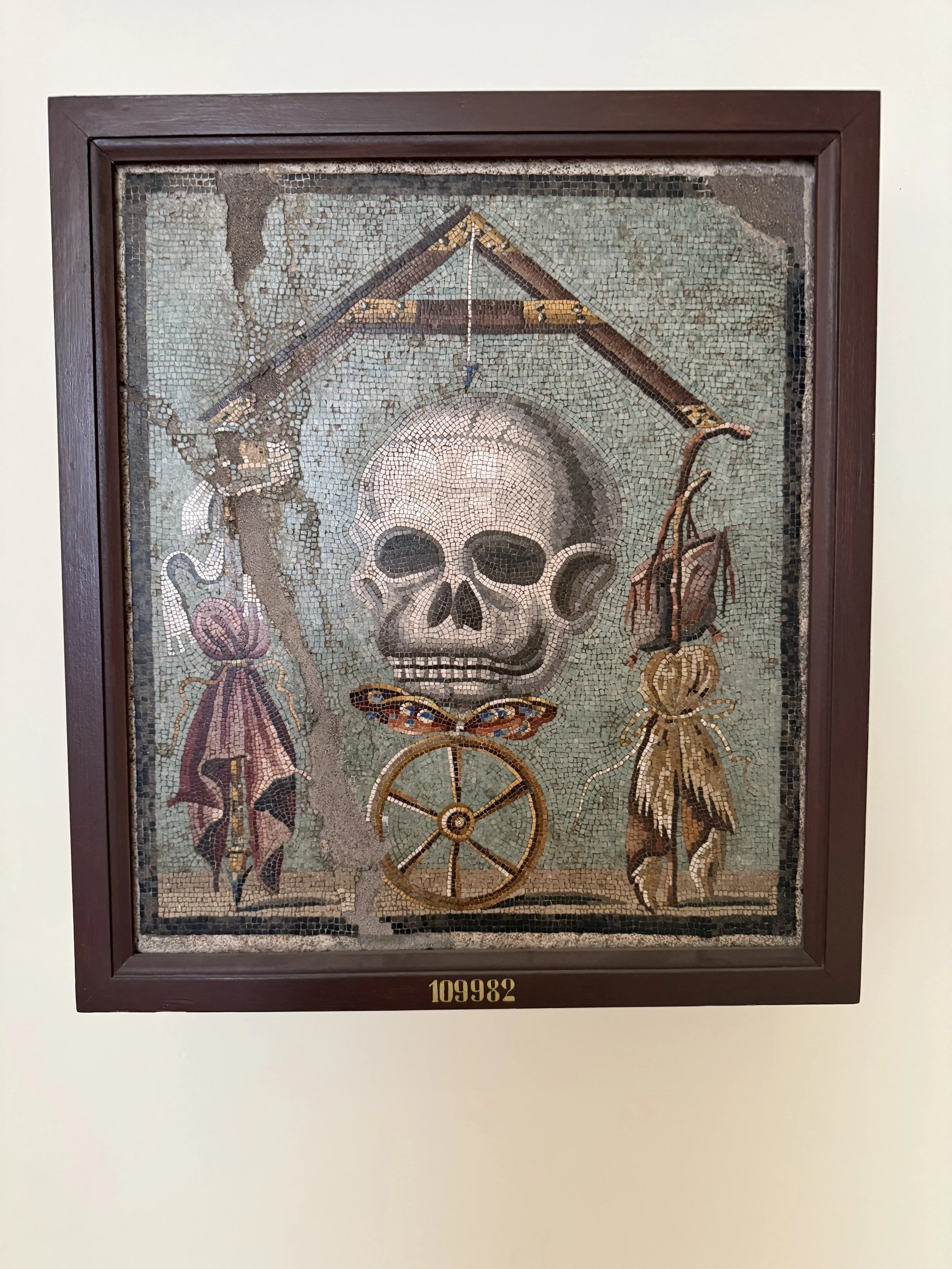 Ancient mosaic art featuring a human skull on top of a chariot wheel, surrounded by floral and animal motifs, framed in dark wood, with a golden number "109982" at the bottom.
