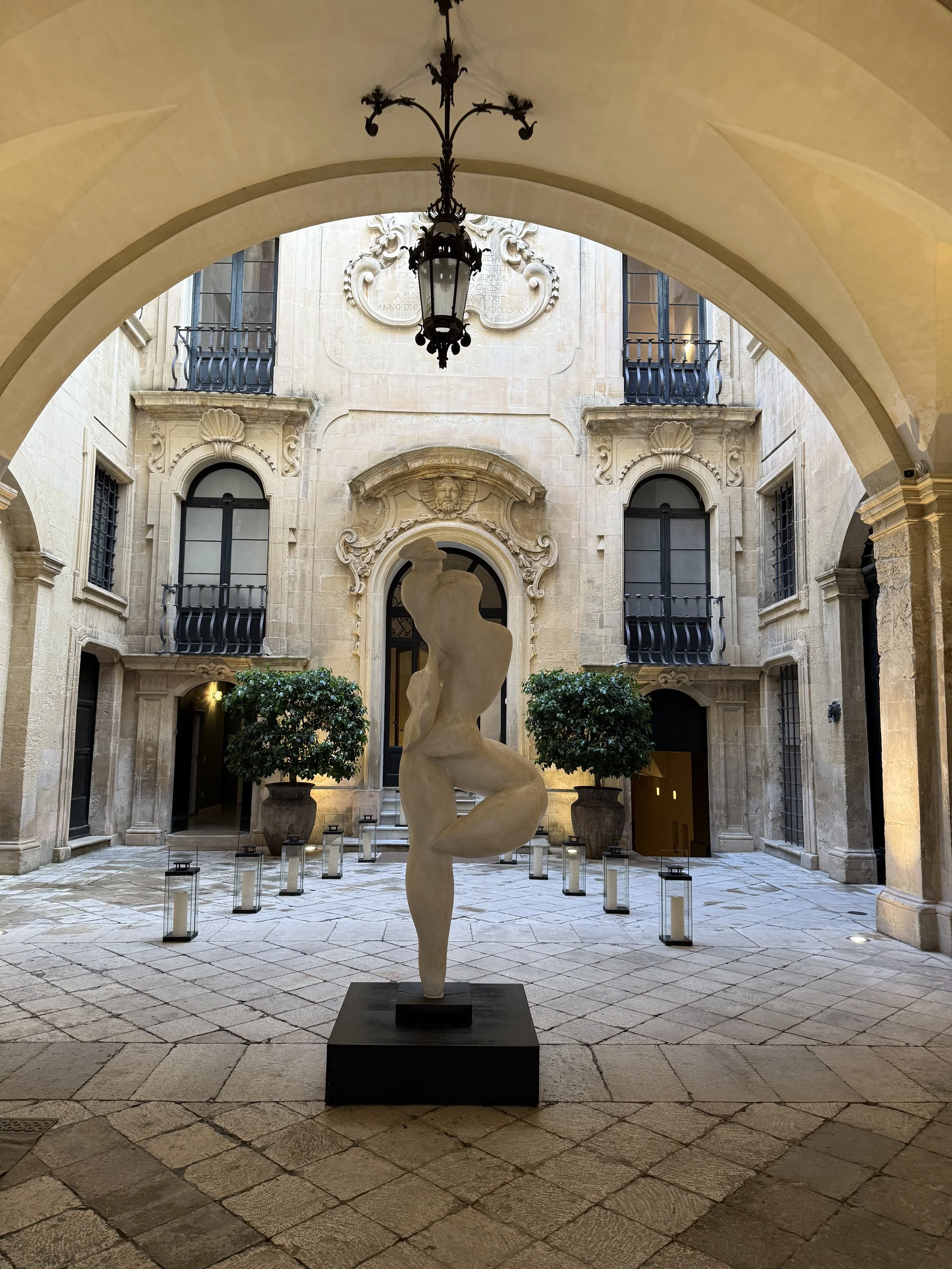 An inner courtyard with a modern abstract statue of a human figure on a black pedestal, surrounded by potted trees and lanterns, with classical architecture details and a decorative stone wall in the background.