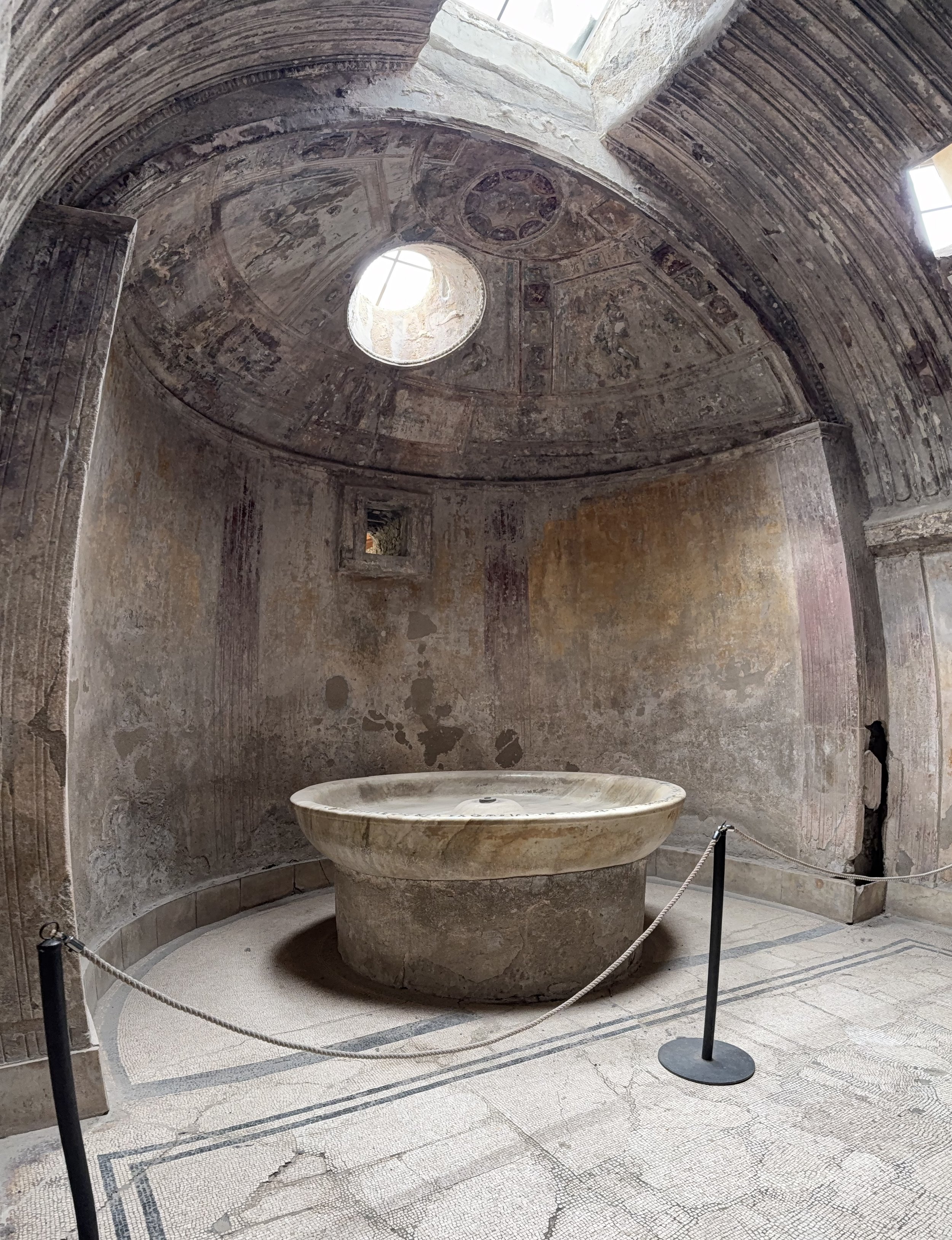 Ancient Roman bathhouse with a circular marble basin in the center, surrounded by stone walls and a mosaic floor with crack patterns, and an oculus window letting light into the space.