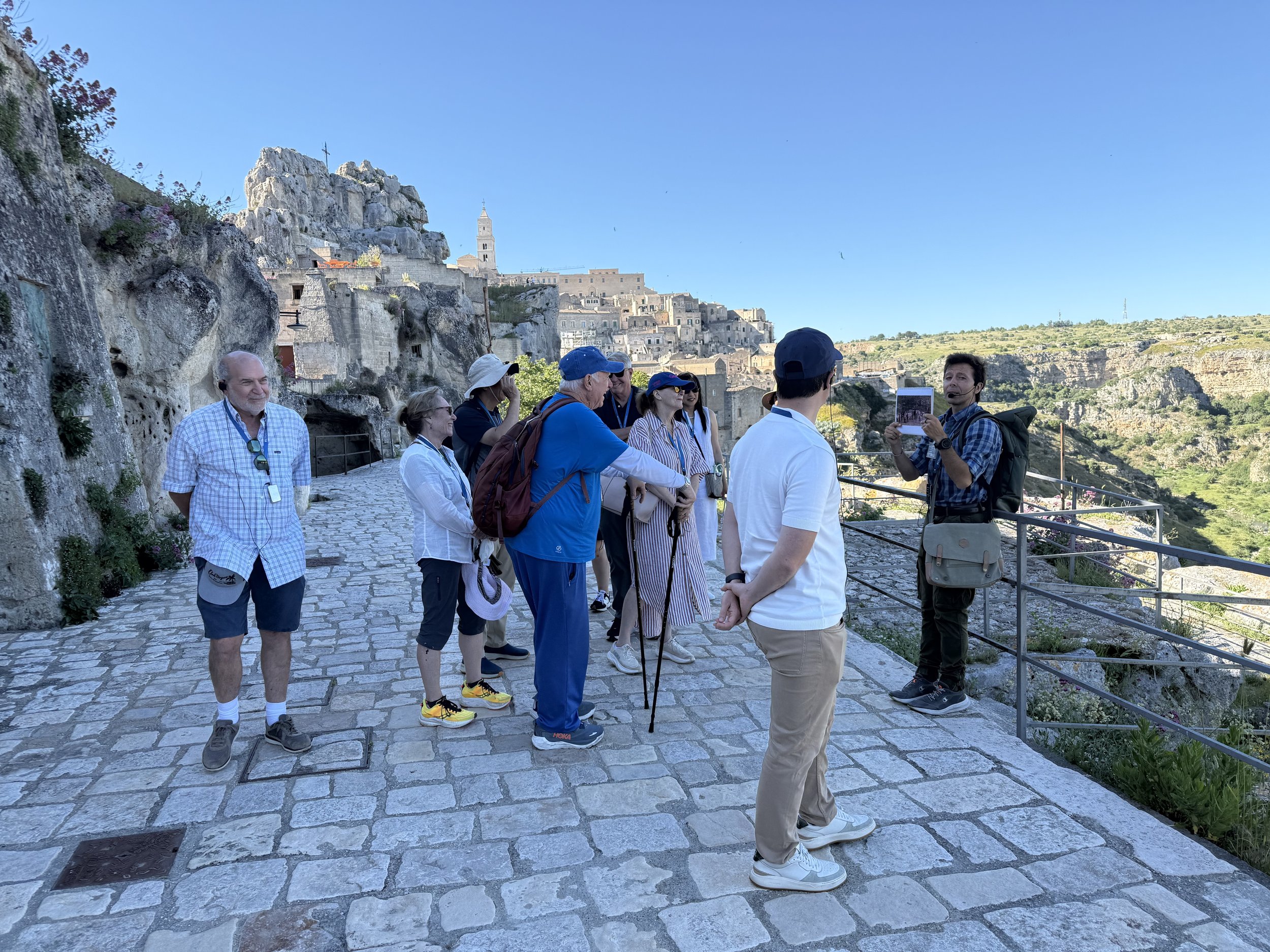 Group of tourists at a hillside overlook, listening to a guide holding a photo, with ancient hillside buildings and a church steeple in the background during daytime.