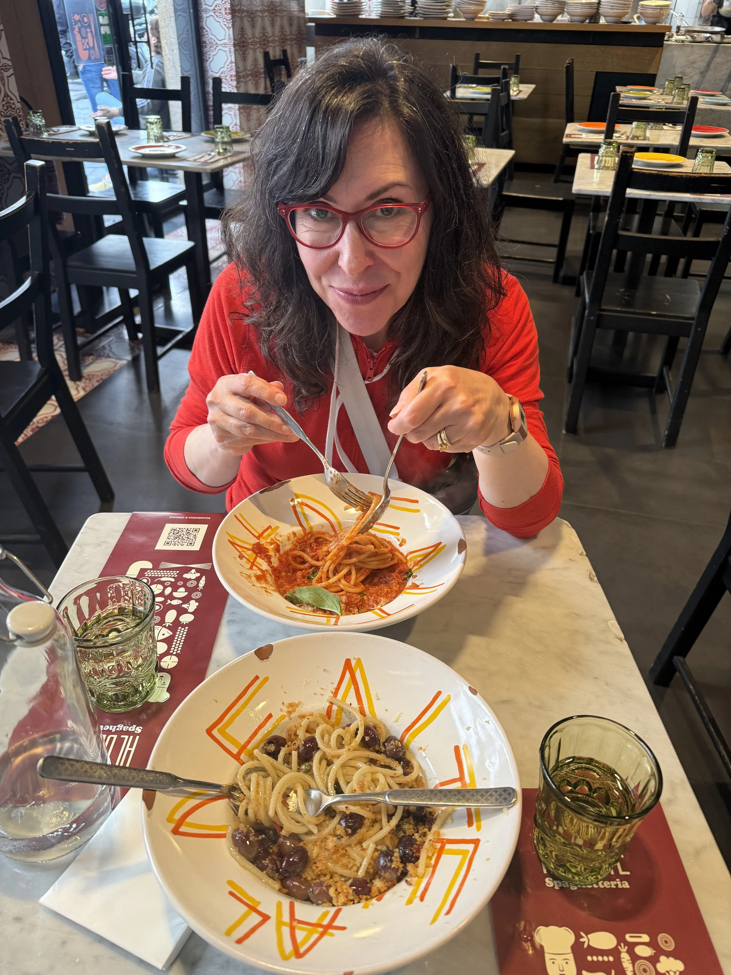 A woman with glasses and curly dark hair, wearing a red top, sitting at a restaurant table, holding a fork and spoon, with bowls of pasta and glasses of water on the table.