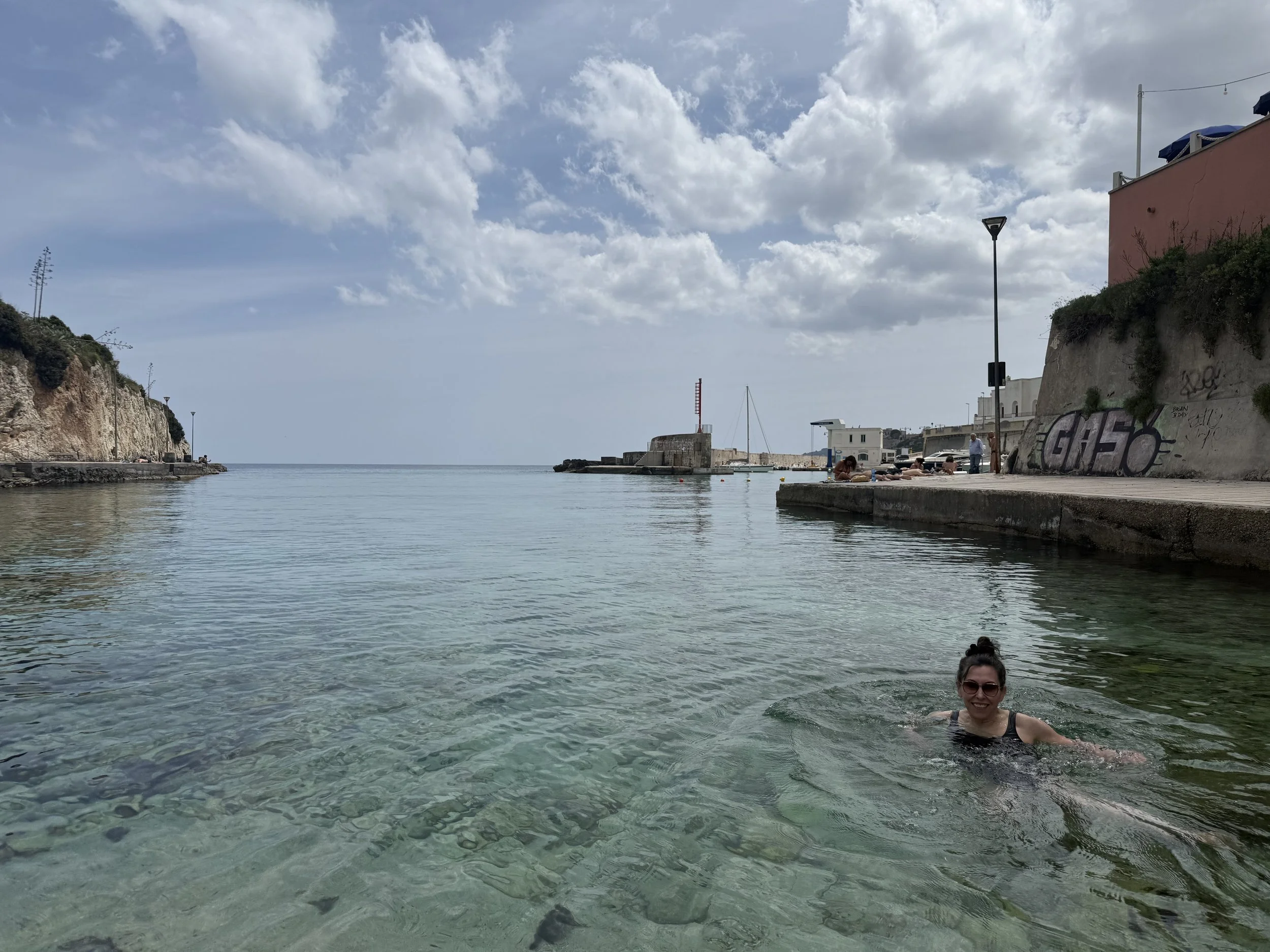A woman with sunglasses smiling while swimming in clear, shallow water near a rocky coastal area with a pink building, lampposts, and other people in the background under a partly cloudy sky.