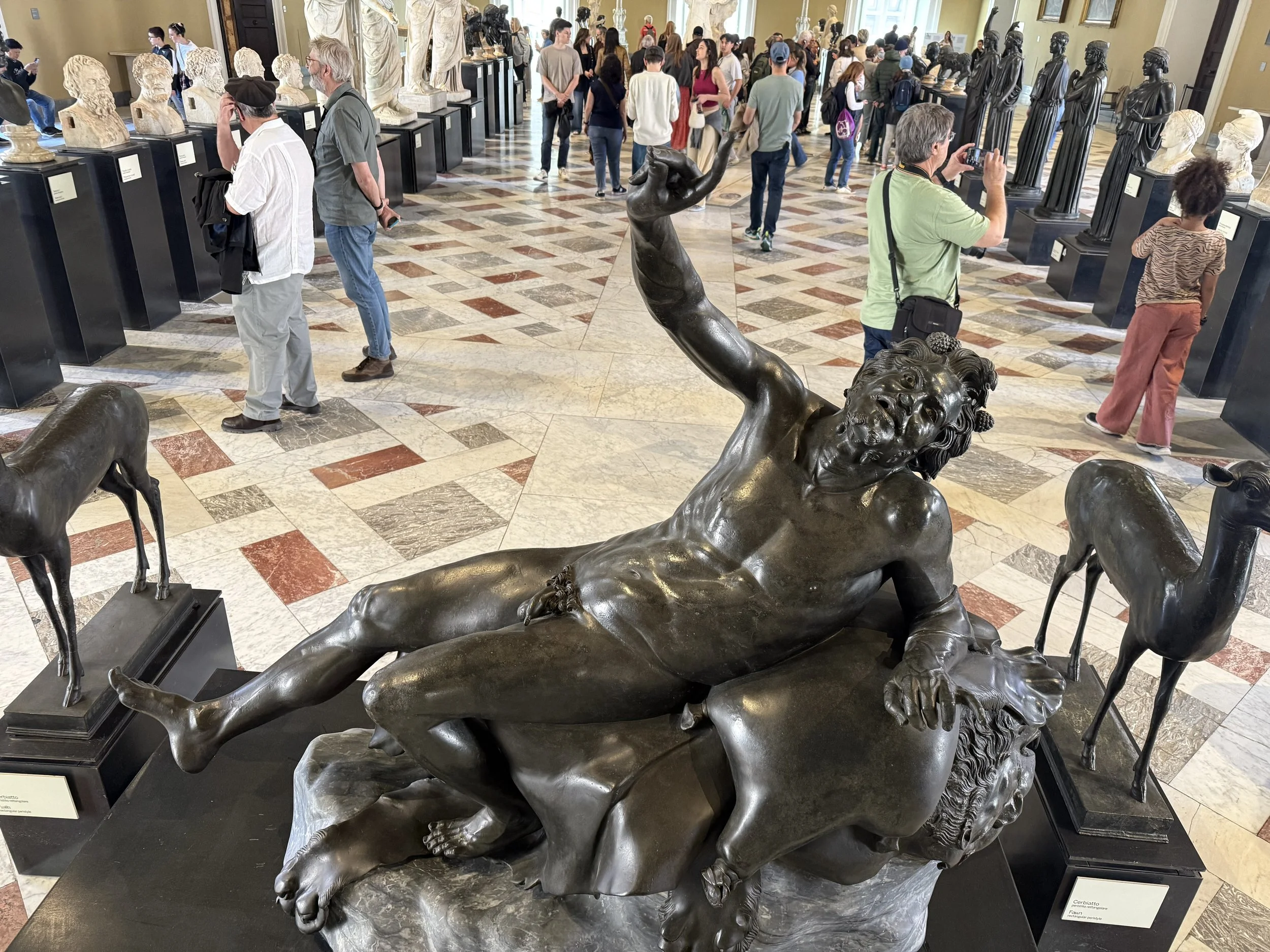Museum gallery with various sculptures and busts, visitors observing and taking photos, marble tiled floor, and a bronze sculpture of a reclining figure in the foreground.
