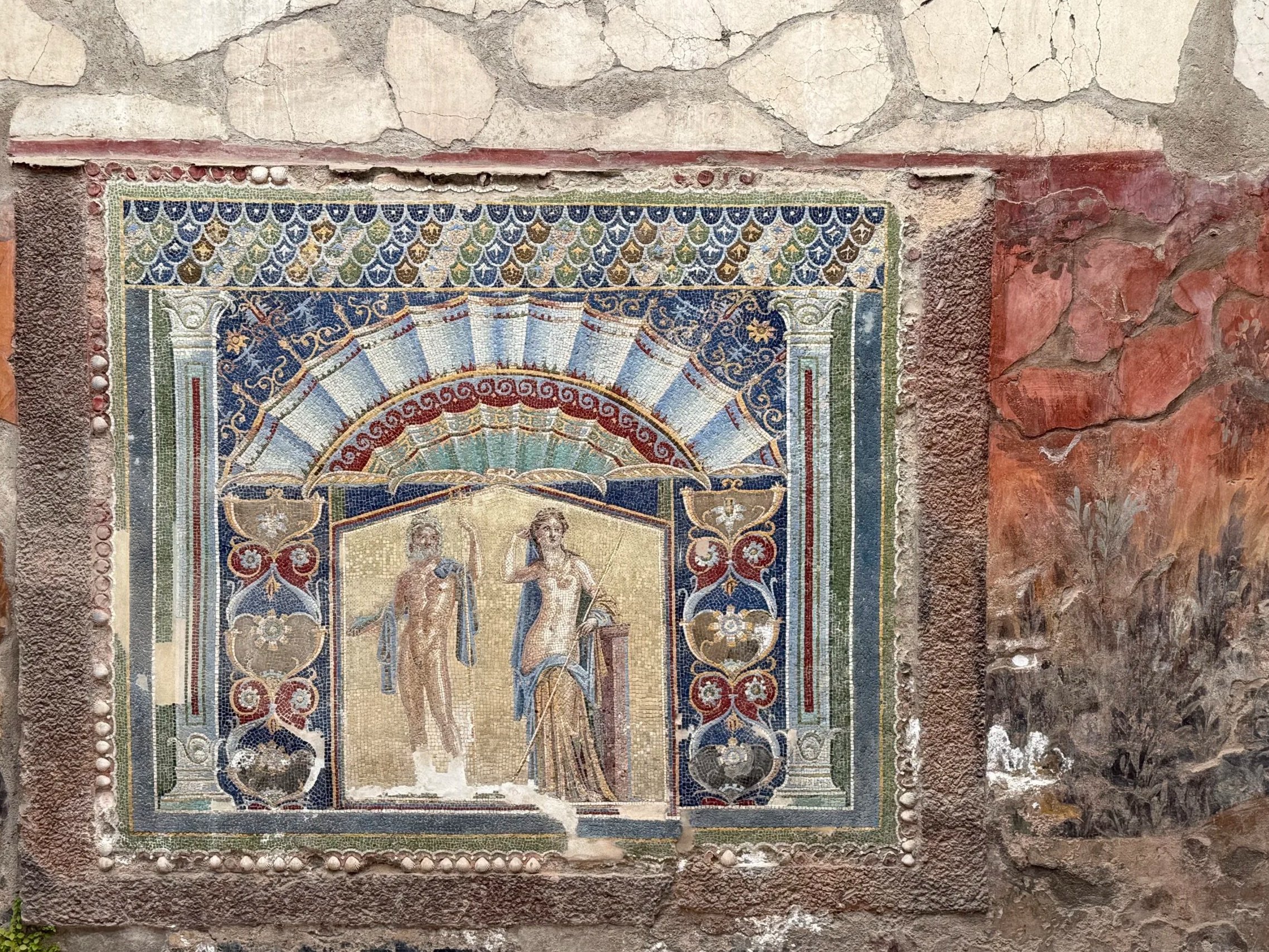 Ancient mosaic artwork depicting two figures, one holding a staff, surrounded by intricate patterns, set in a weathered stone wall with visible cracks and mortar.