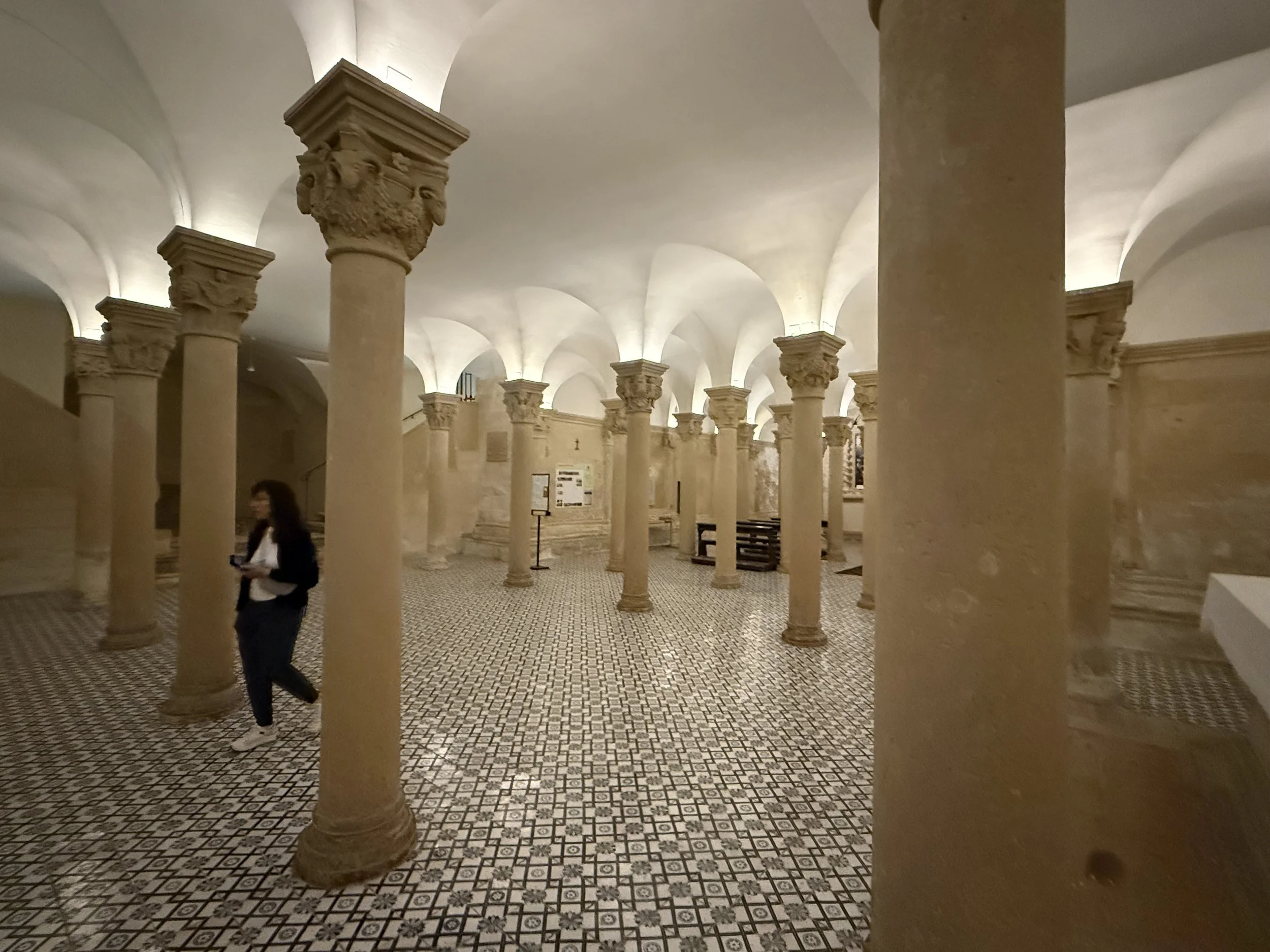 Interior of a historical building with numerous stone columns supporting a vaulted ceiling, illuminated with soft lighting, and patterned tile flooring; a woman in a dark suit and a man in a white shirt and dark jacket are walking inside.