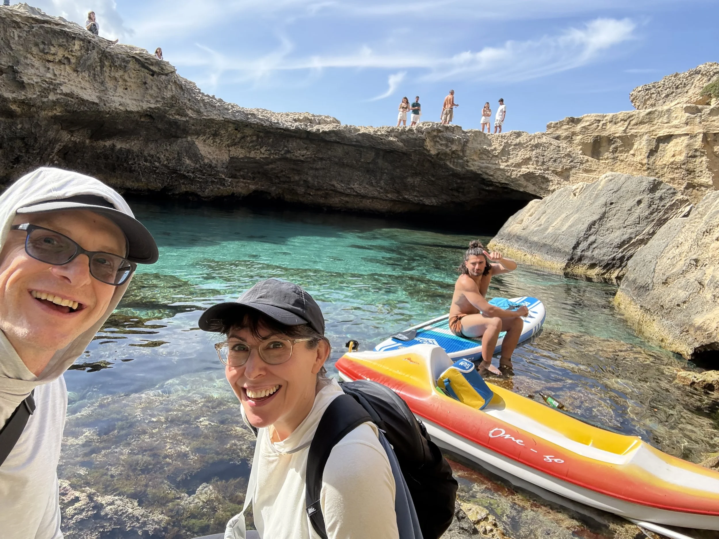 A group of people at a rocky beach with clear turquoise water, some sitting on a paddleboard, others standing on the rocks with a blue sky in the background. One person in the foreground is smiling at the camera.