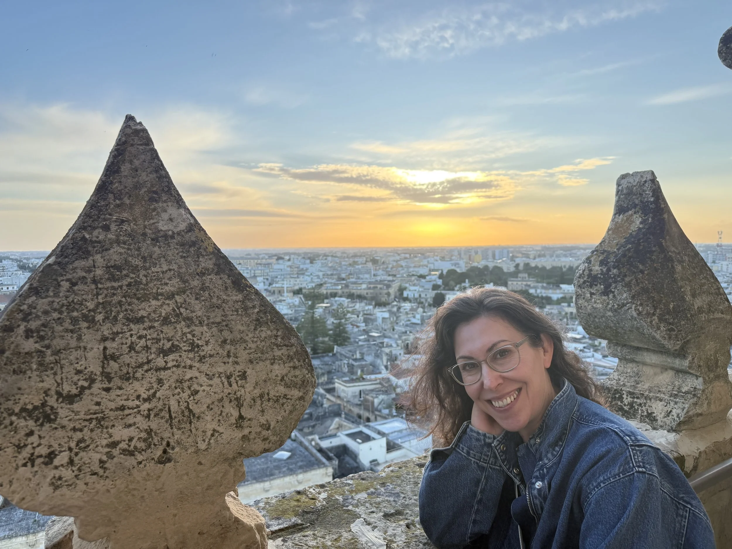 A woman smiling, wearing glasses and a denim jacket, posing between two large, weathered stones on a rooftop at sunset with city buildings in the background.