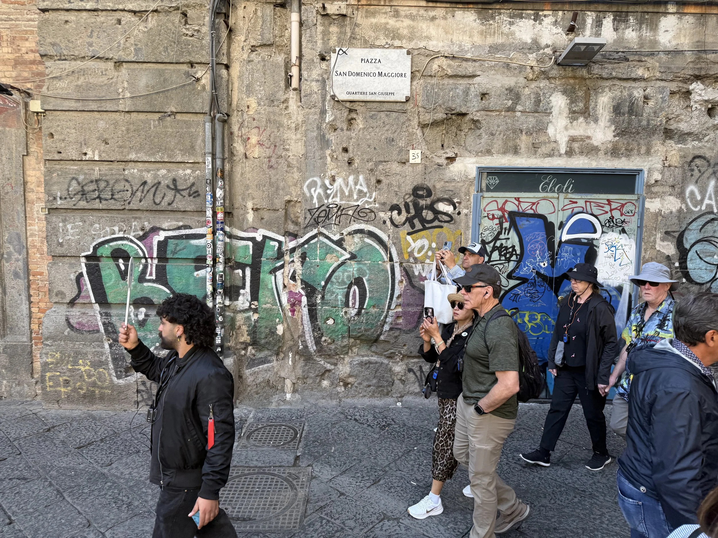 A group of people walking on a city street with graffiti-covered walls in the background. The wall features a sign indicating a piazza called San Domenico Maggiore in the San Giuseppe neighborhood.