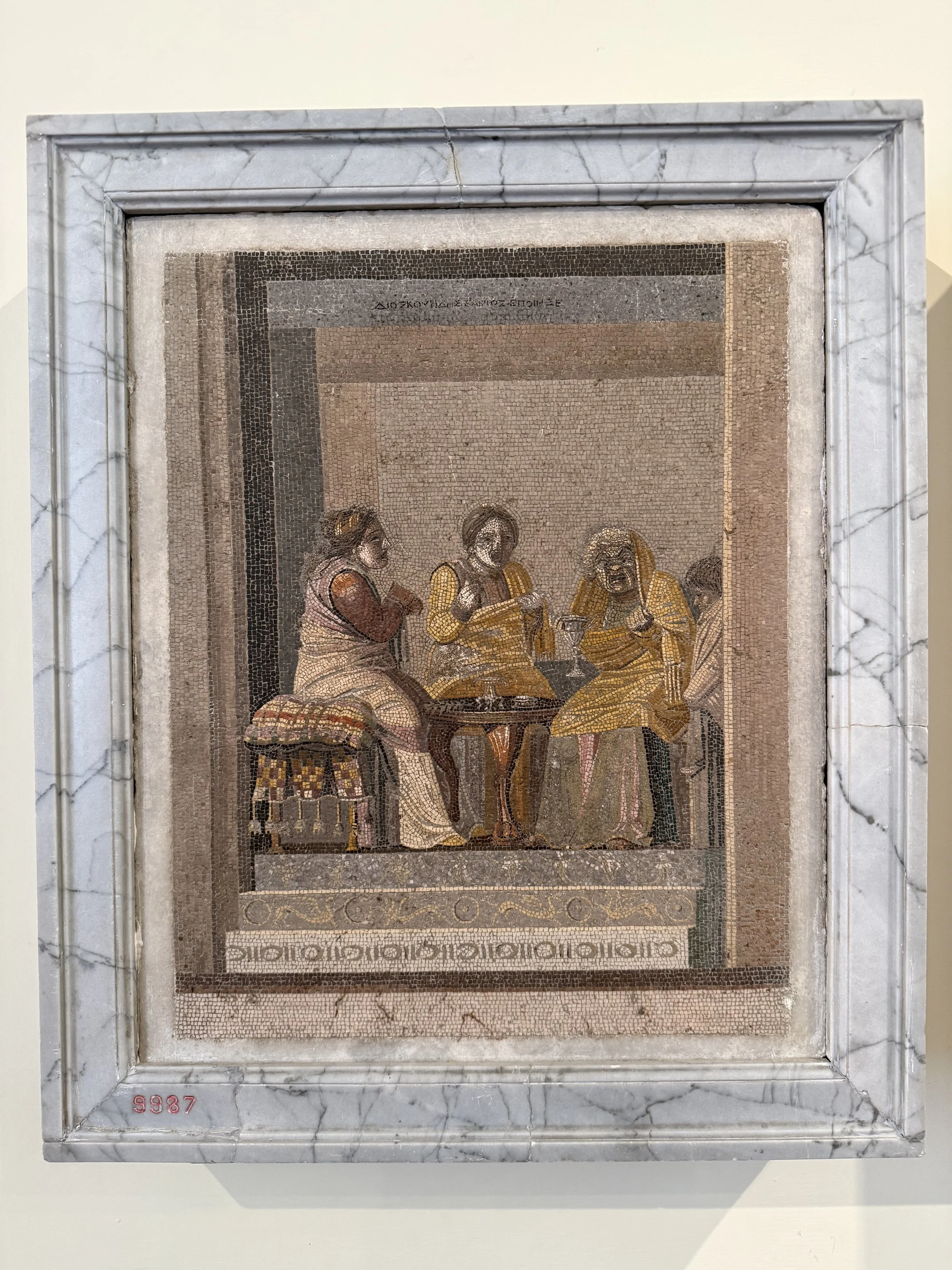 A mosaic artwork depicting three women in traditional clothing, engaged in a discussion or sharing a moment. The mosaic is framed with a marble border and is displayed on a wall.