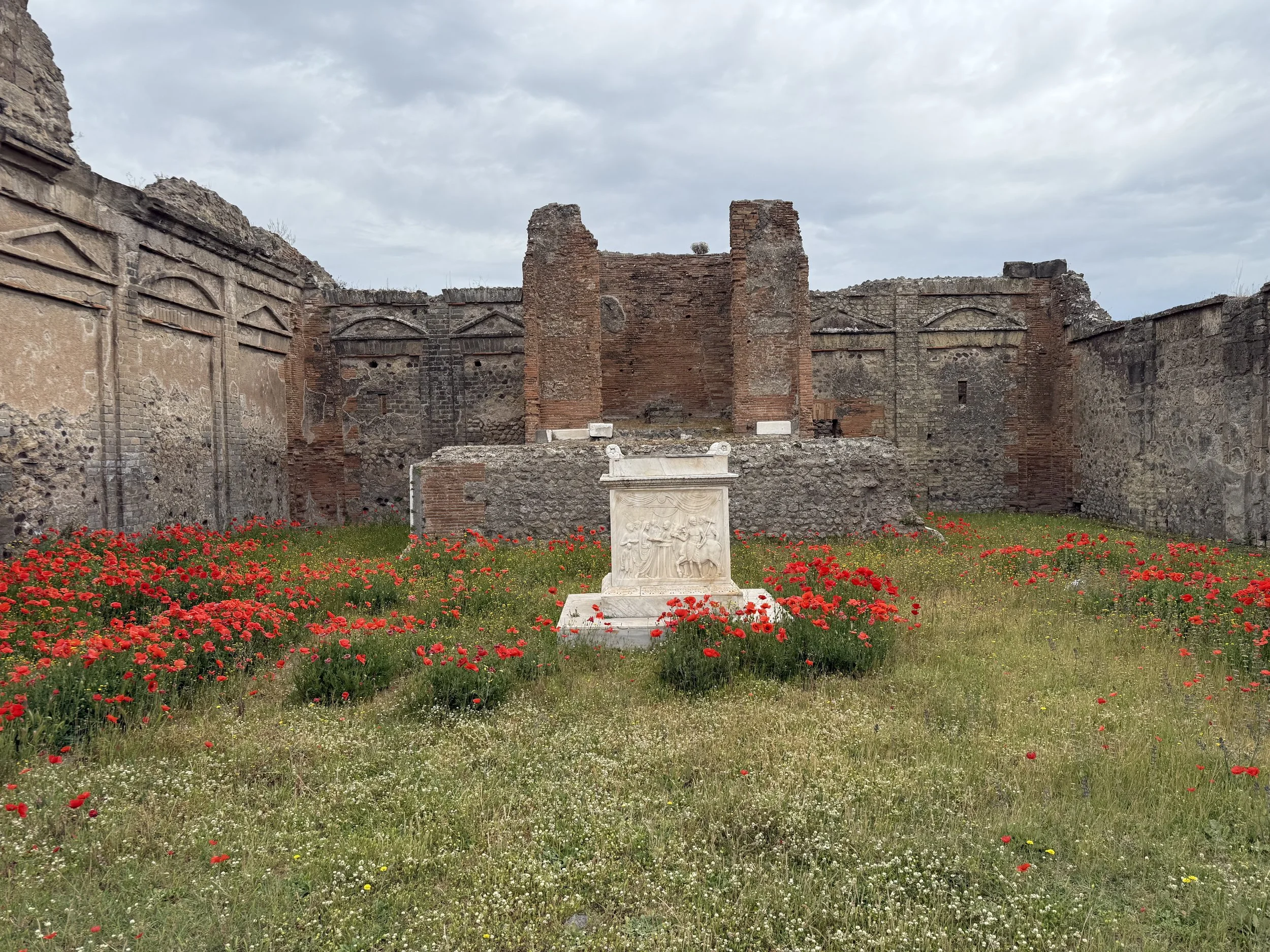 Ruins of an ancient stone structure with a small white marble sculpture in front, surrounded by red poppies and green grass, under a cloudy sky.