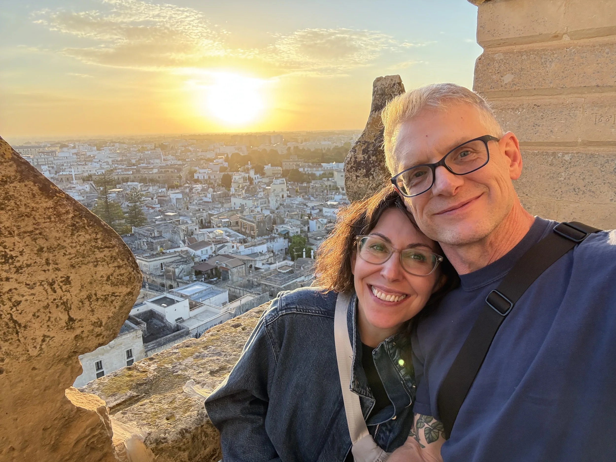 A man with gray hair and glasses and a woman with long brown hair and glasses are smiling and taking a selfie on a rooftop at sunset, with a city view in the background.