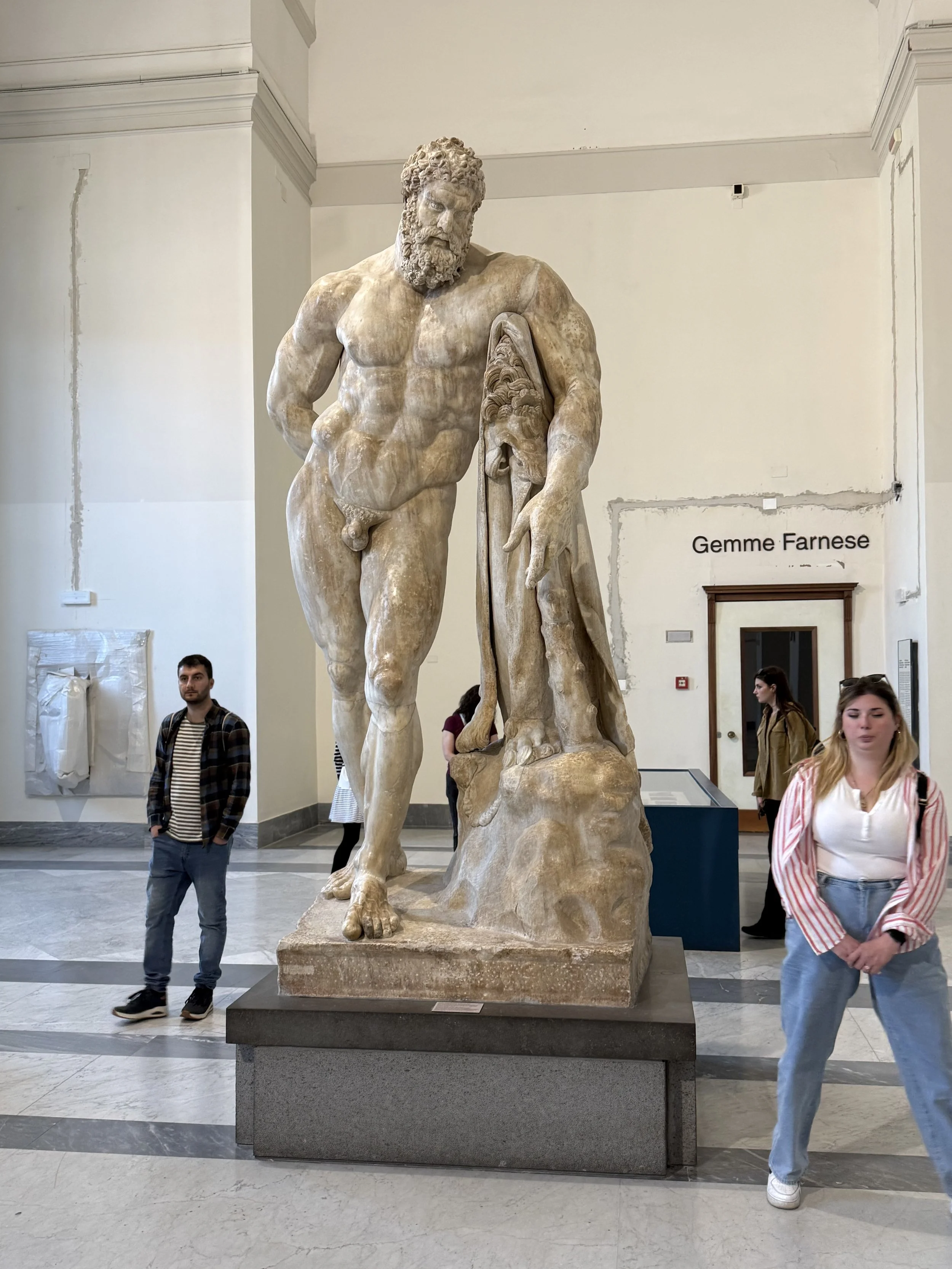 Statue of a muscular bearded man with a cloth draped over his left arm, standing on a stone pedestal in a museum, with visitors walking by.