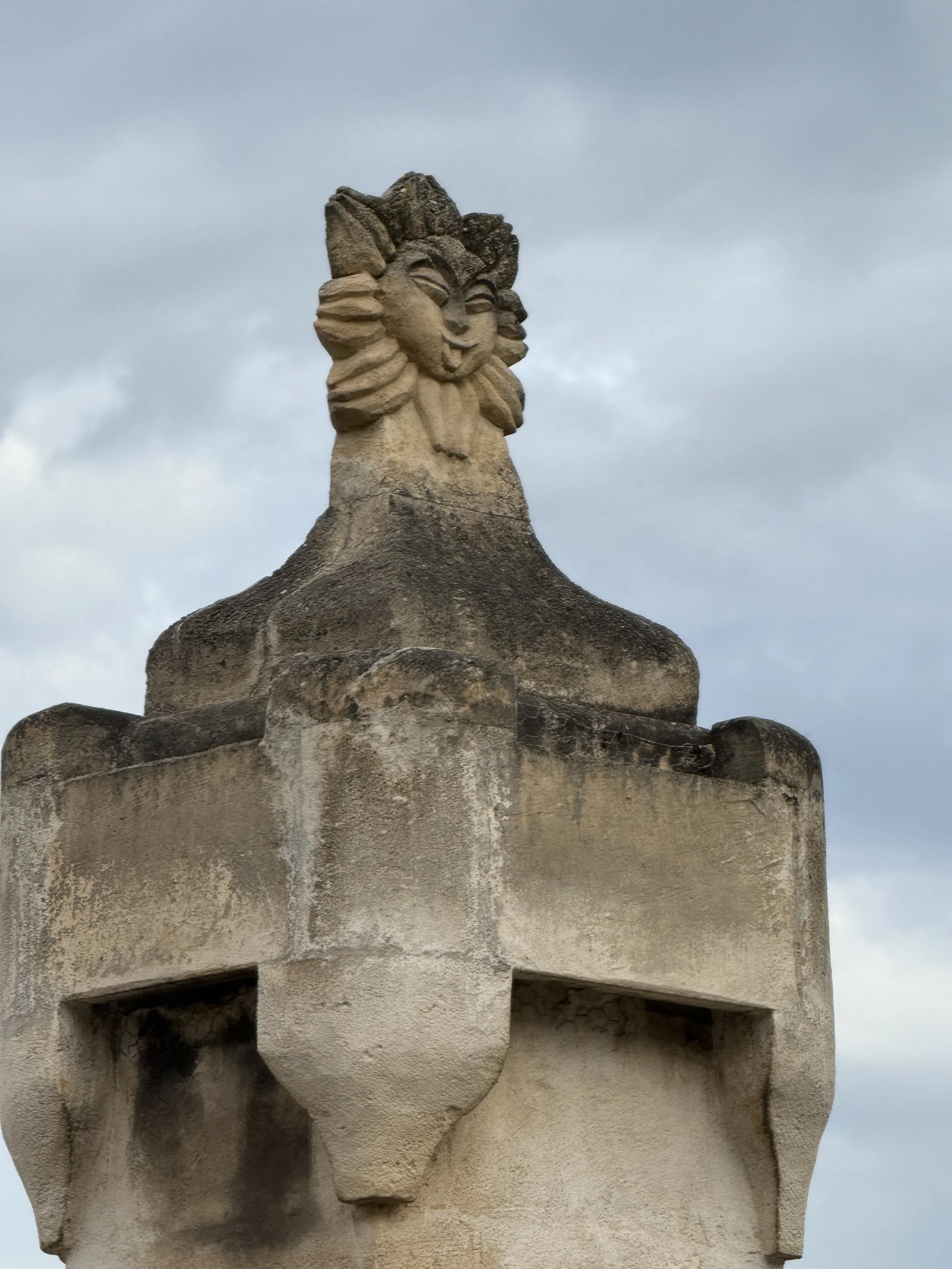 Close-up of a stone sculpture depicting a smiling human face with stylized hair, part of a larger architectural structure against a cloudy sky.