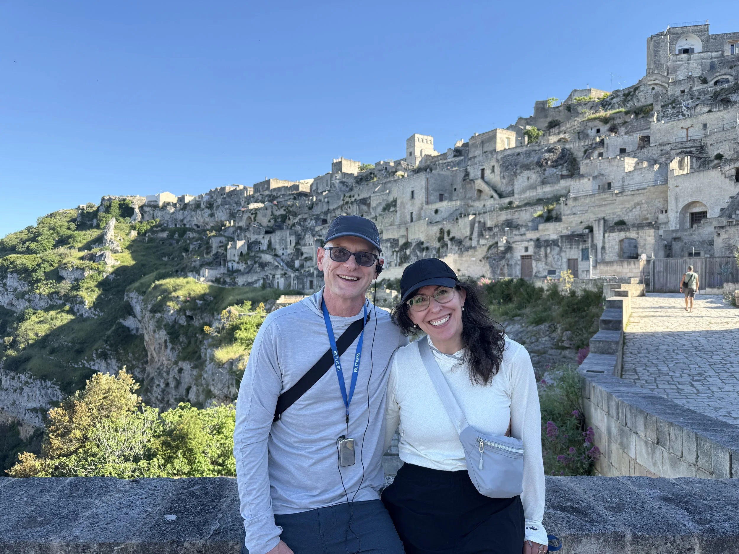 Two happy women posing for a photo in front of an ancient stone hillside city with structures built into the hillside, under a clear blue sky.