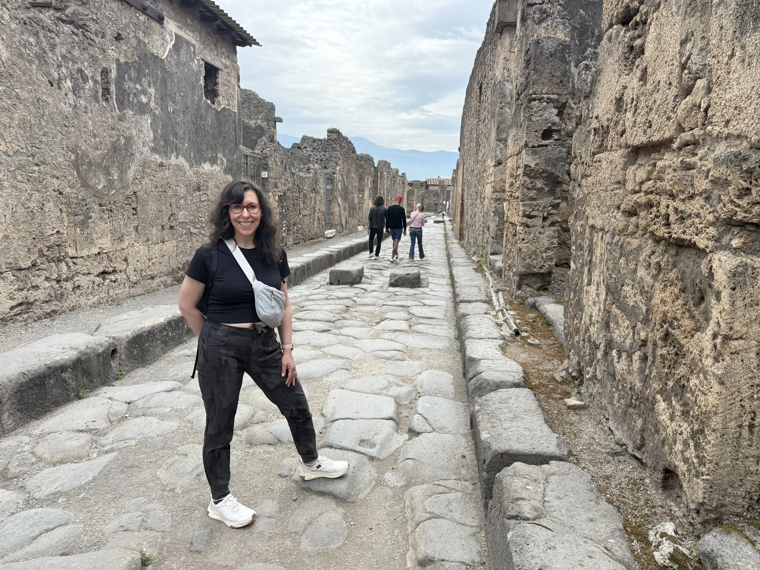 A smiling woman with long dark hair and red glasses standing on an ancient cobblestone street with stone ruins on either side and a mountain in the background.