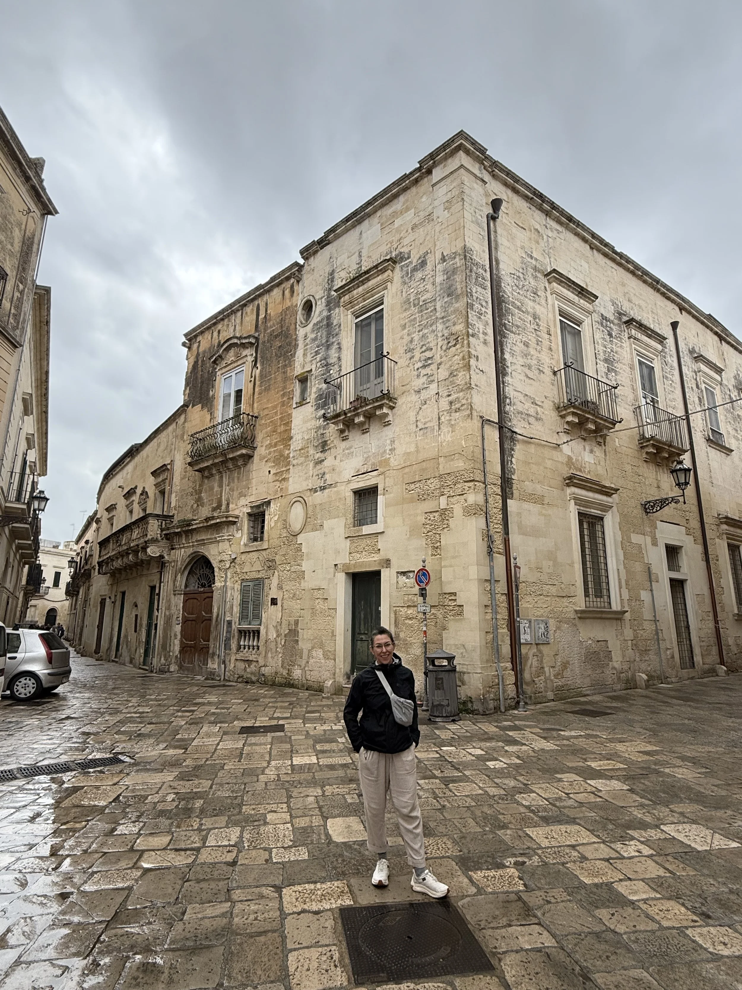 A woman standing on a cobblestone street in front of an old stone building with windows and small balconies, under a cloudy sky, wearing a black jacket, beige pants, and white sneakers.