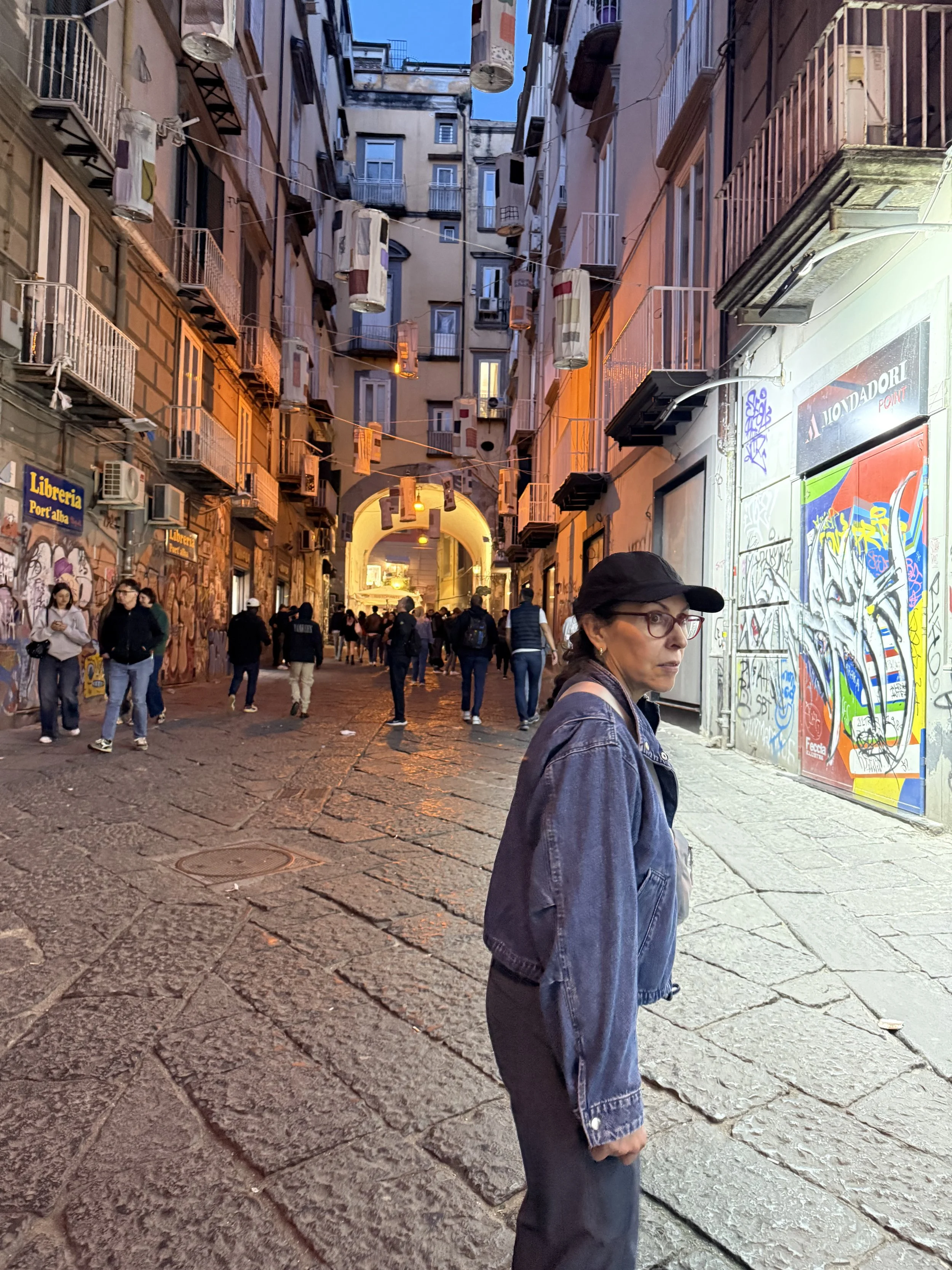 Street scene in an urban area with a crowd of people walking on cobblestone pavement. Tall buildings with graffiti, storefronts, and hanging laundry or decorations between the buildings. A woman in a black cap and denim jacket is in the foreground.
