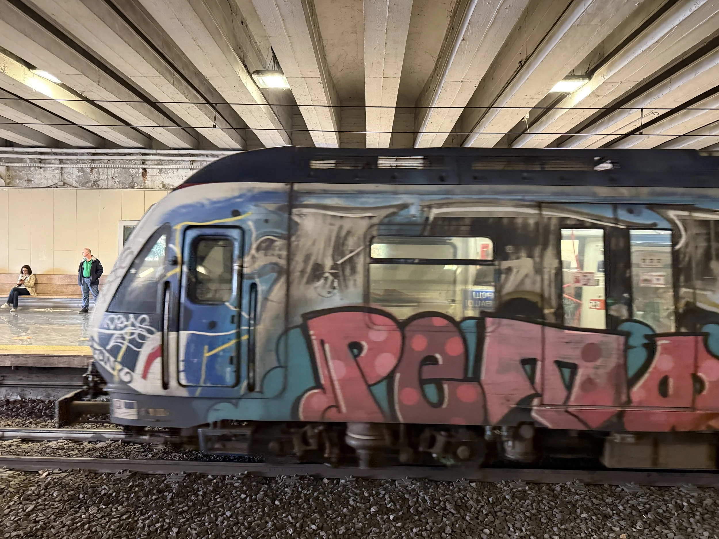 A subway train with graffiti on its side traveling through a station with concrete ceiling and passengers waiting on the platform.