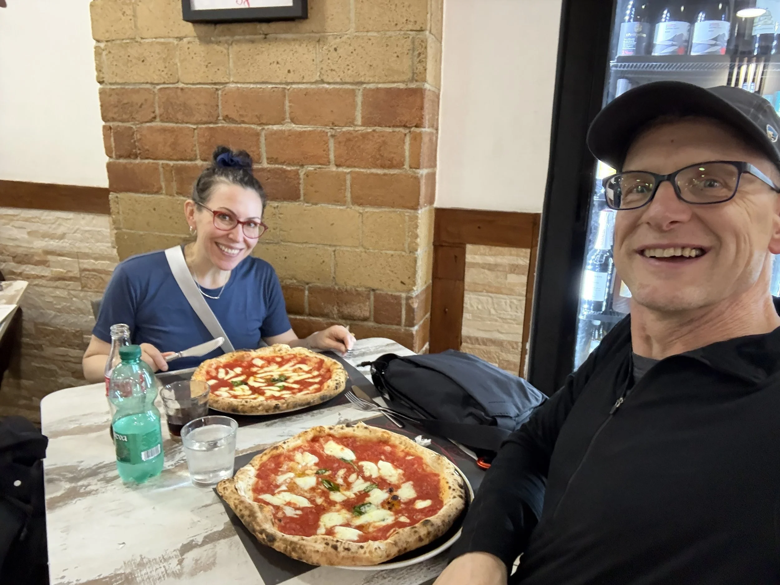 A man and a woman sitting at a table in a restaurant with two pizzas, drinks, and water bottles. The woman has glasses, red hair, and a blue shirt. The man has glasses, a cap, and is smiling.