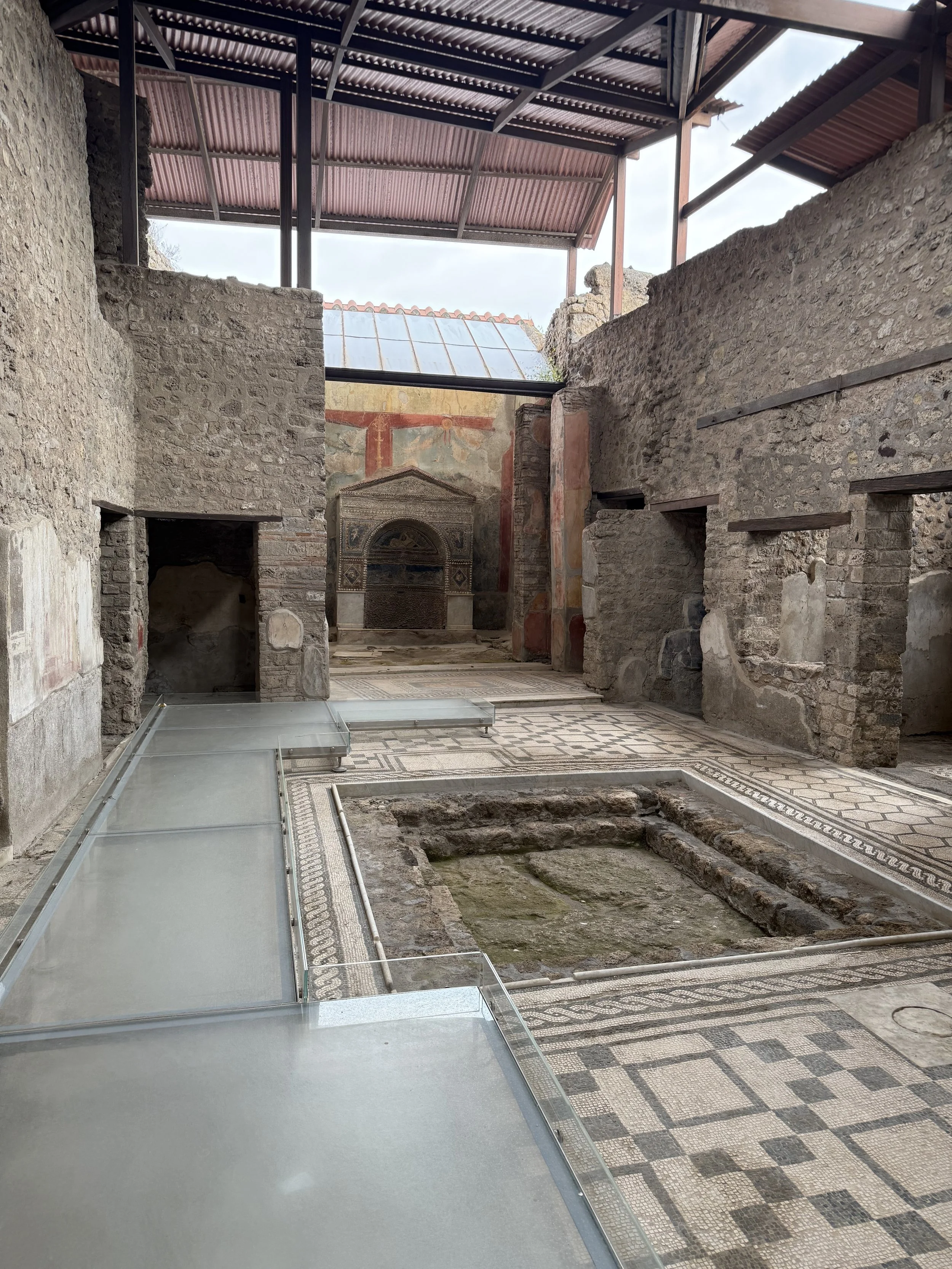 Ancient Roman archaeological site with partially reconstructed walls, mosaic flooring, and a vaulted frescoed wall at the back, with modern glass and metal structural supports.