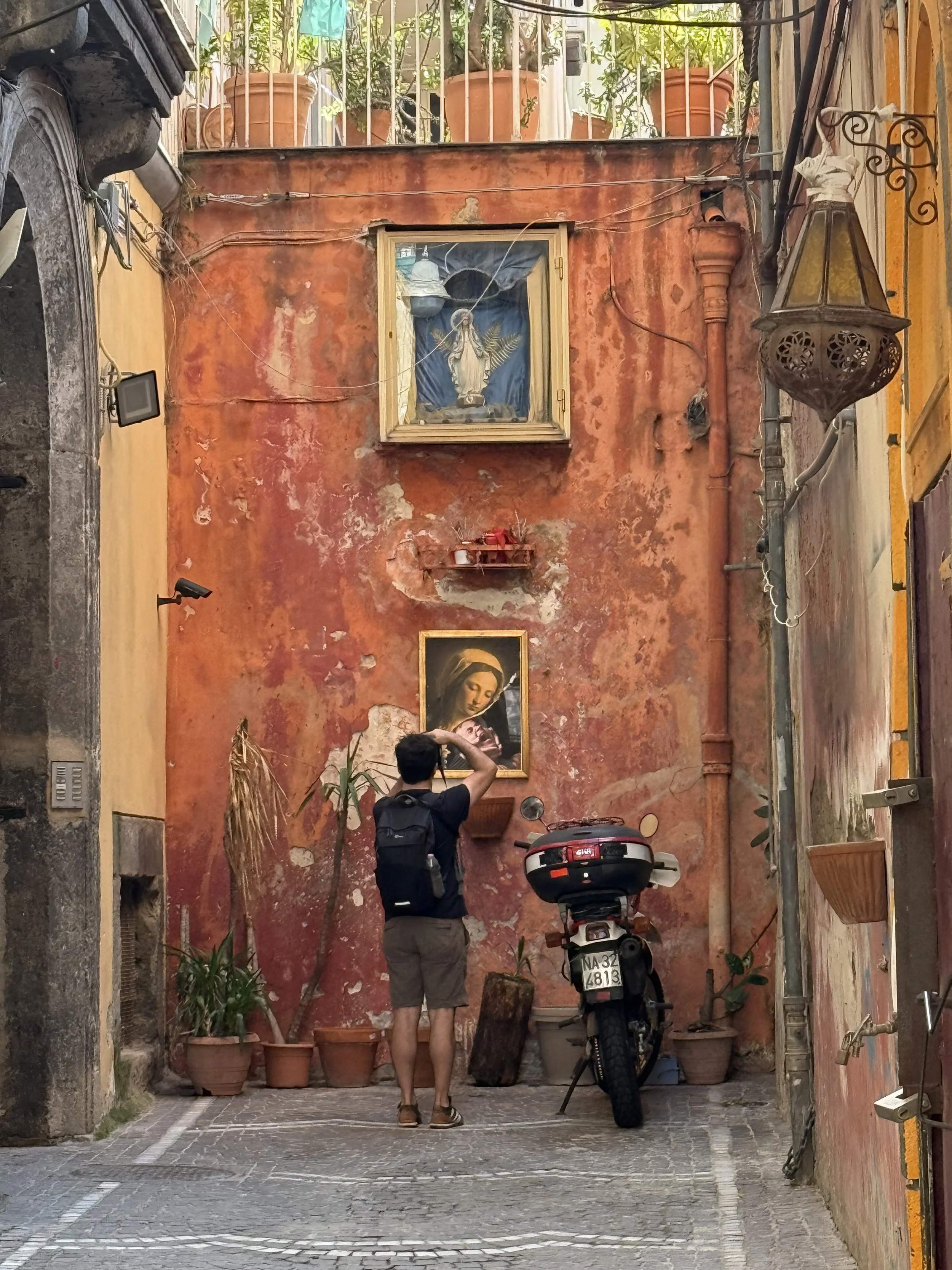 A narrow street with a person taking a photo of a red wall decorated with religious artwork and potted plants. A motorcycle is parked in front of the wall, and another small potted plant is on the ground to the left.