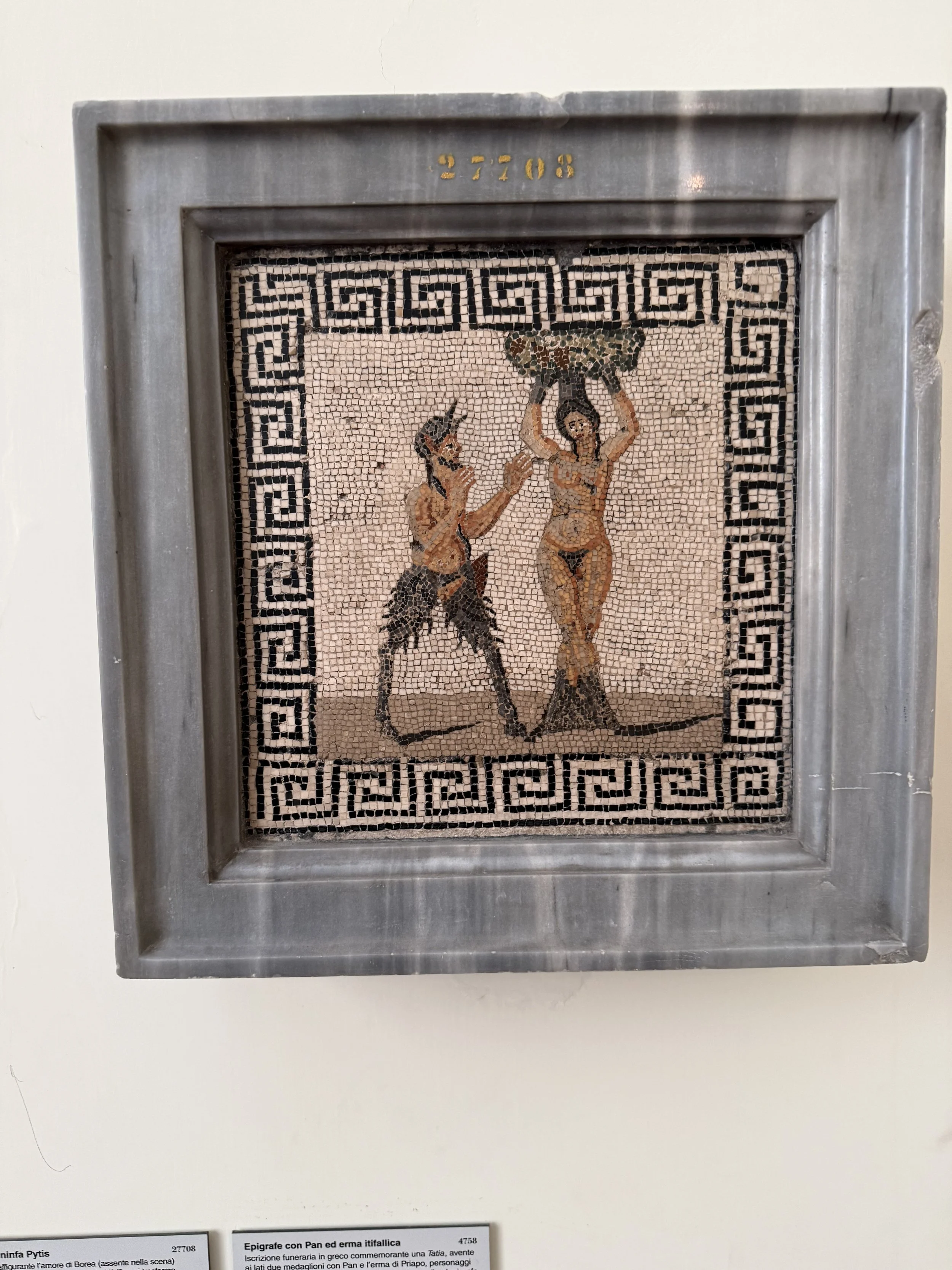 Ancient mosaic artwork depicting two figures, one with a bow and arrow and the other holding a bowl or vessel, surrounded by a Greek key border, displayed behind a metal frame.