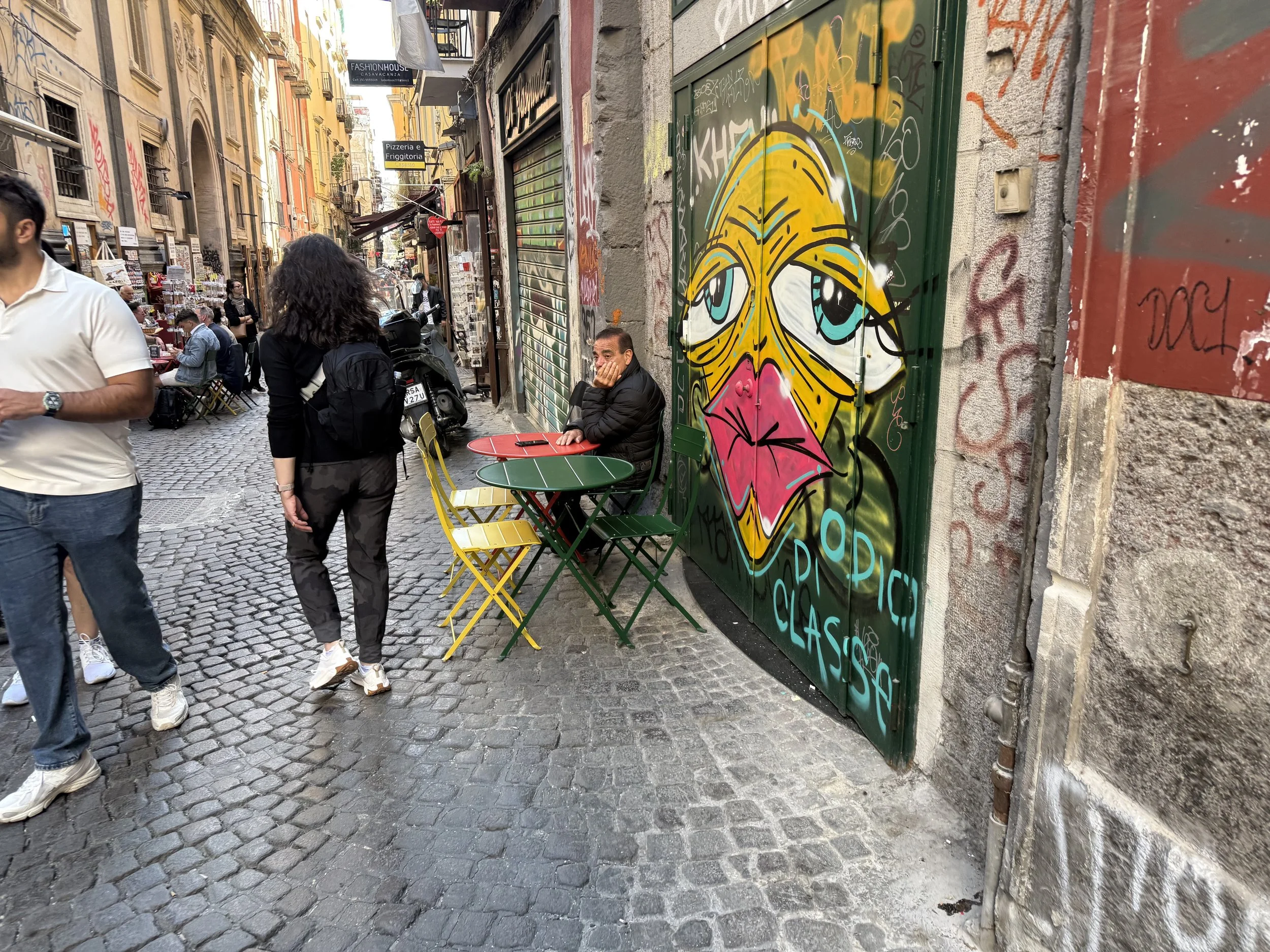A street scene with graffiti art of a woman's face on a green door, people walking and sitting at outdoor tables, and a man sitting with his face resting on his hand beside the graffiti.
