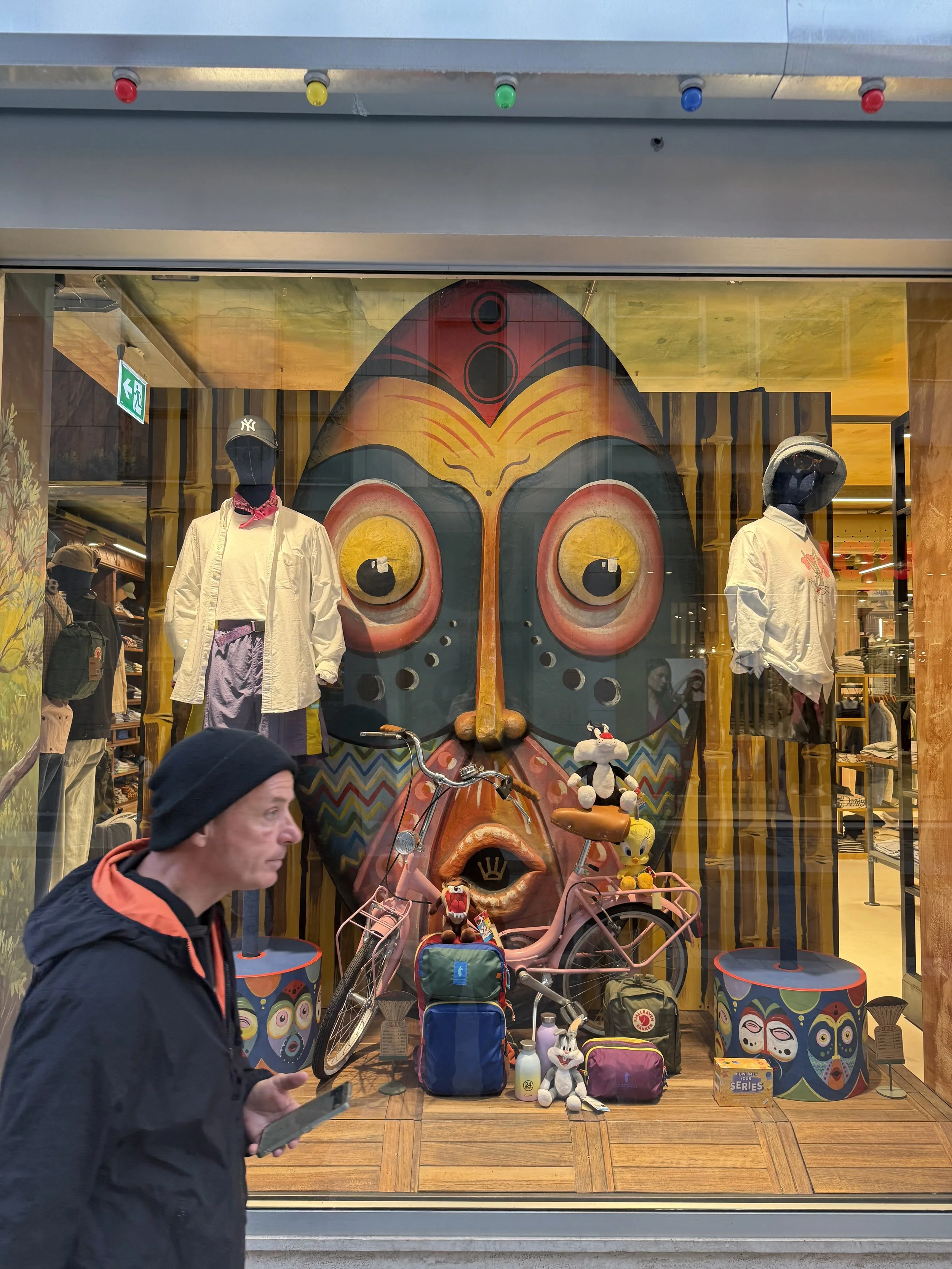 Store window display featuring items such as mannequins dressed in casual clothing, a large butterfly artwork on the back wall, bicycles, plush toys, backpacks, and small tables, with a man walking past holding a smartphone.