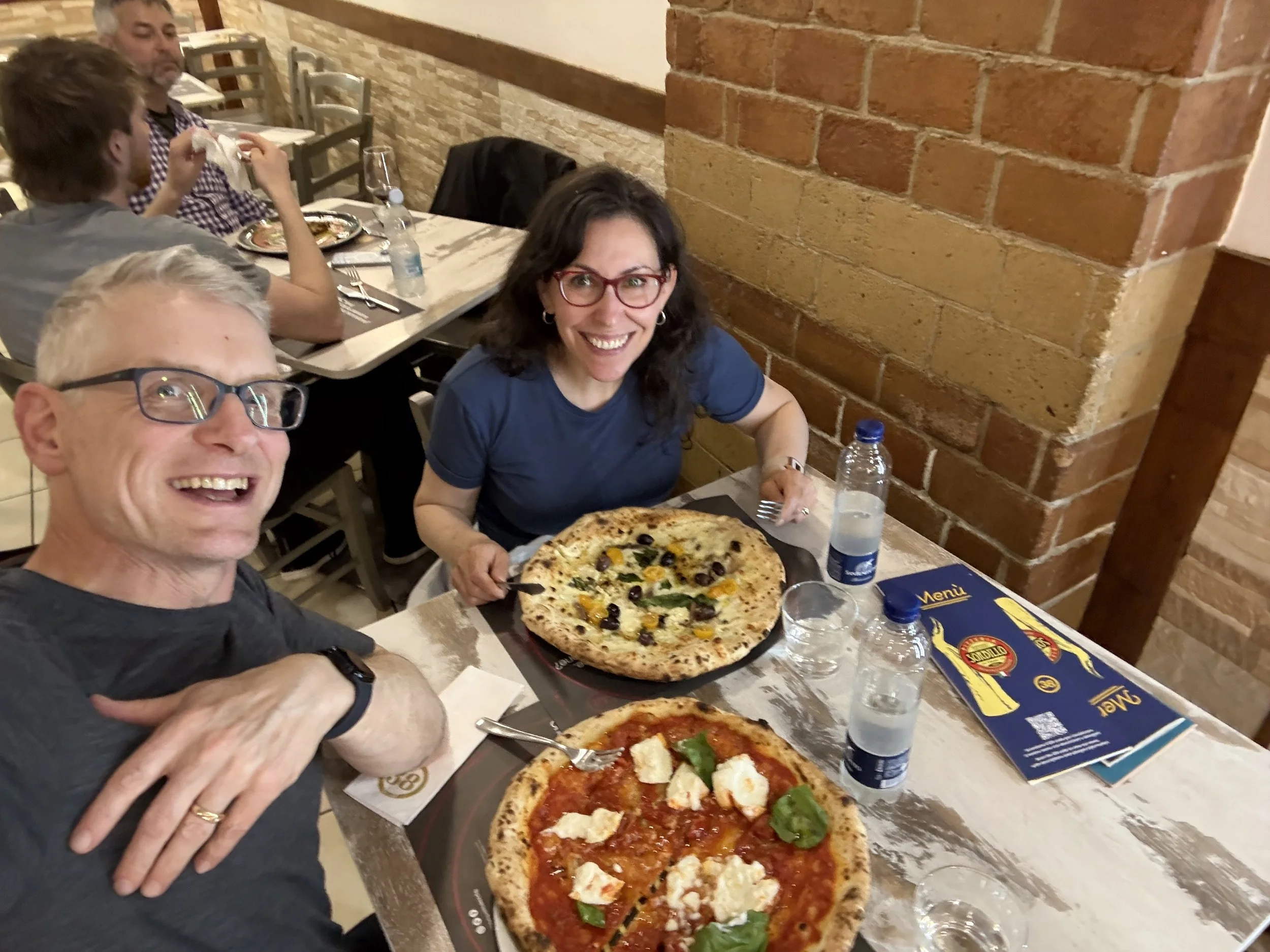 Four people are at a table in a restaurant with brick walls. Two women and two men are smiling and enjoying pizza. One woman with dark hair and glasses appears to be holding a pizza; the man in the foreground with glasses is taking a selfie. There ar