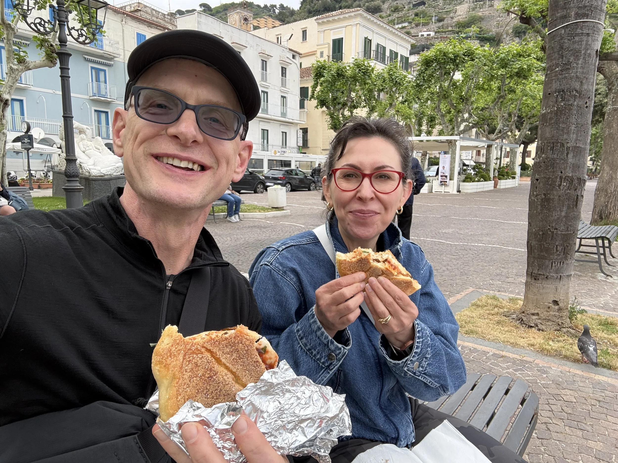 A smiling man wearing glasses and a cap, and a woman with red glasses, both holding sandwiches wrapped in foil, sitting outdoors in a town square with buildings, trees, and a lamppost in the background.