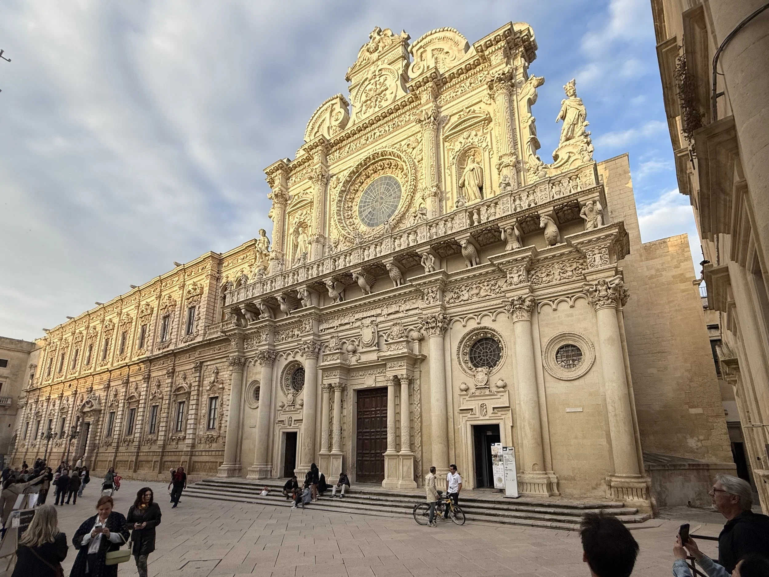Historical cathedral with ornate architecture, sculptures, and a rose window façade, crowds of people in front, bright sky with clouds.