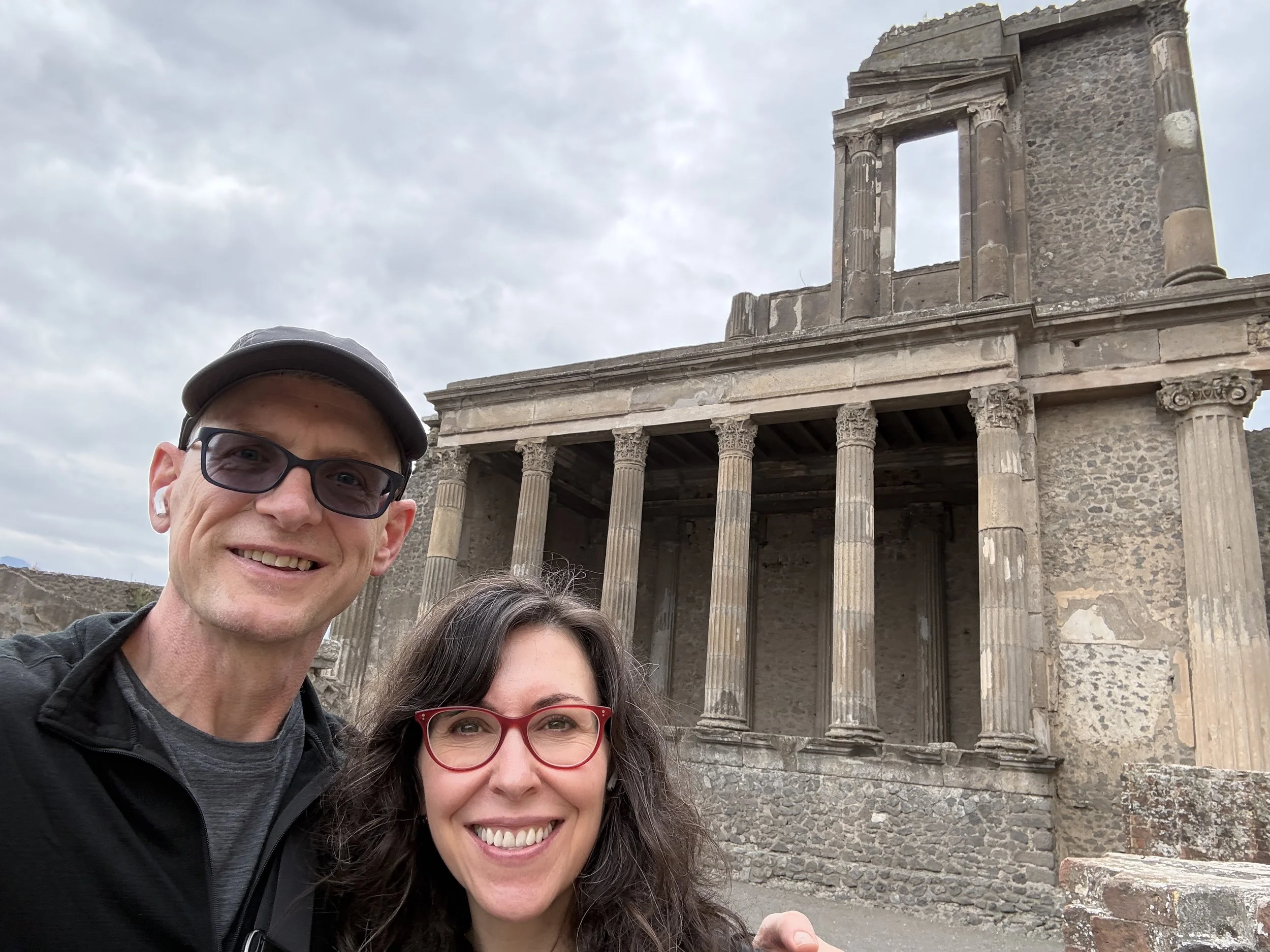 A smiling man and woman taking a selfie in front of ancient ruins with stone columns and a stone building with a window opening, under cloudy sky.