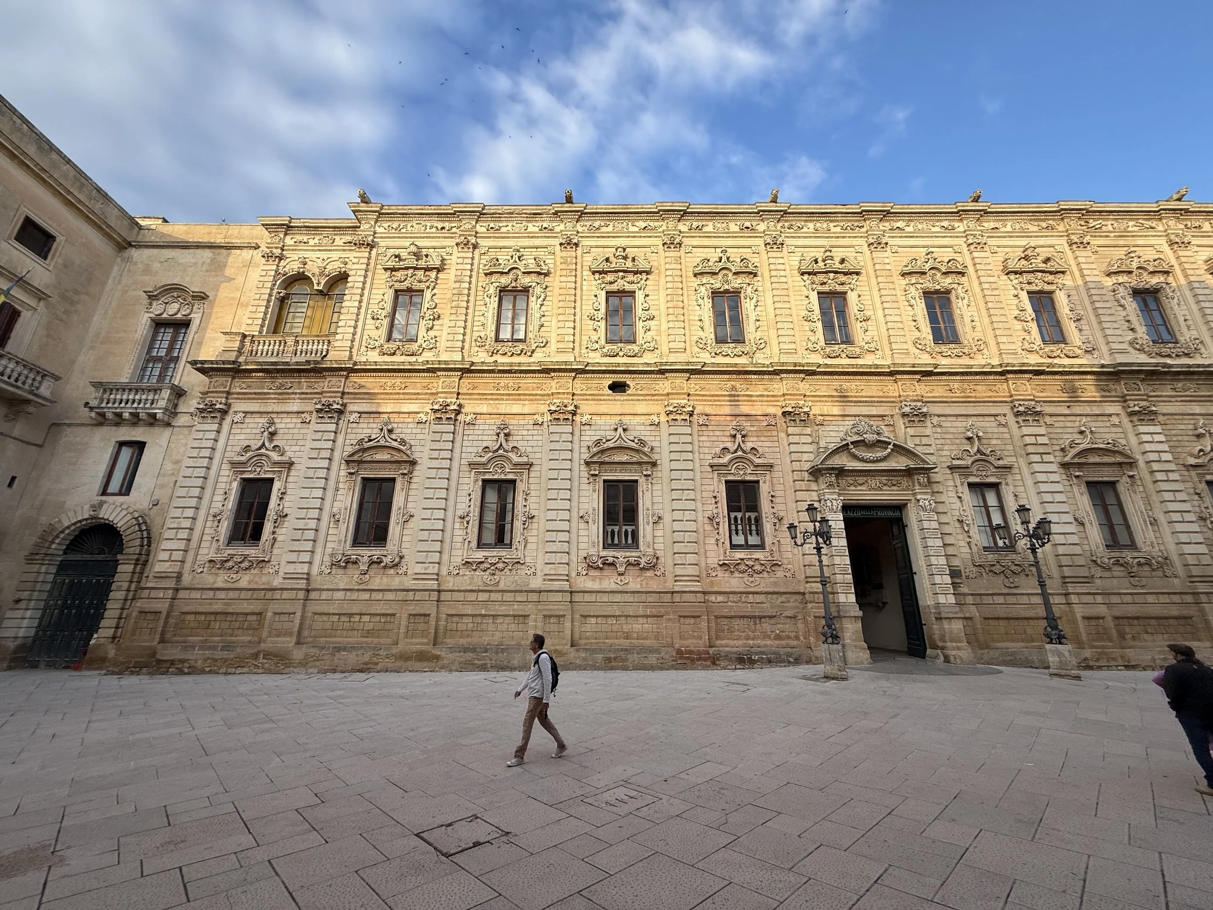 A historic beige stone building with ornate architectural details, arched windows, and decorative moldings. The building is situated on a paved plaza with a few pedestrians walking nearby, against a backdrop of blue sky with some clouds and birds fly