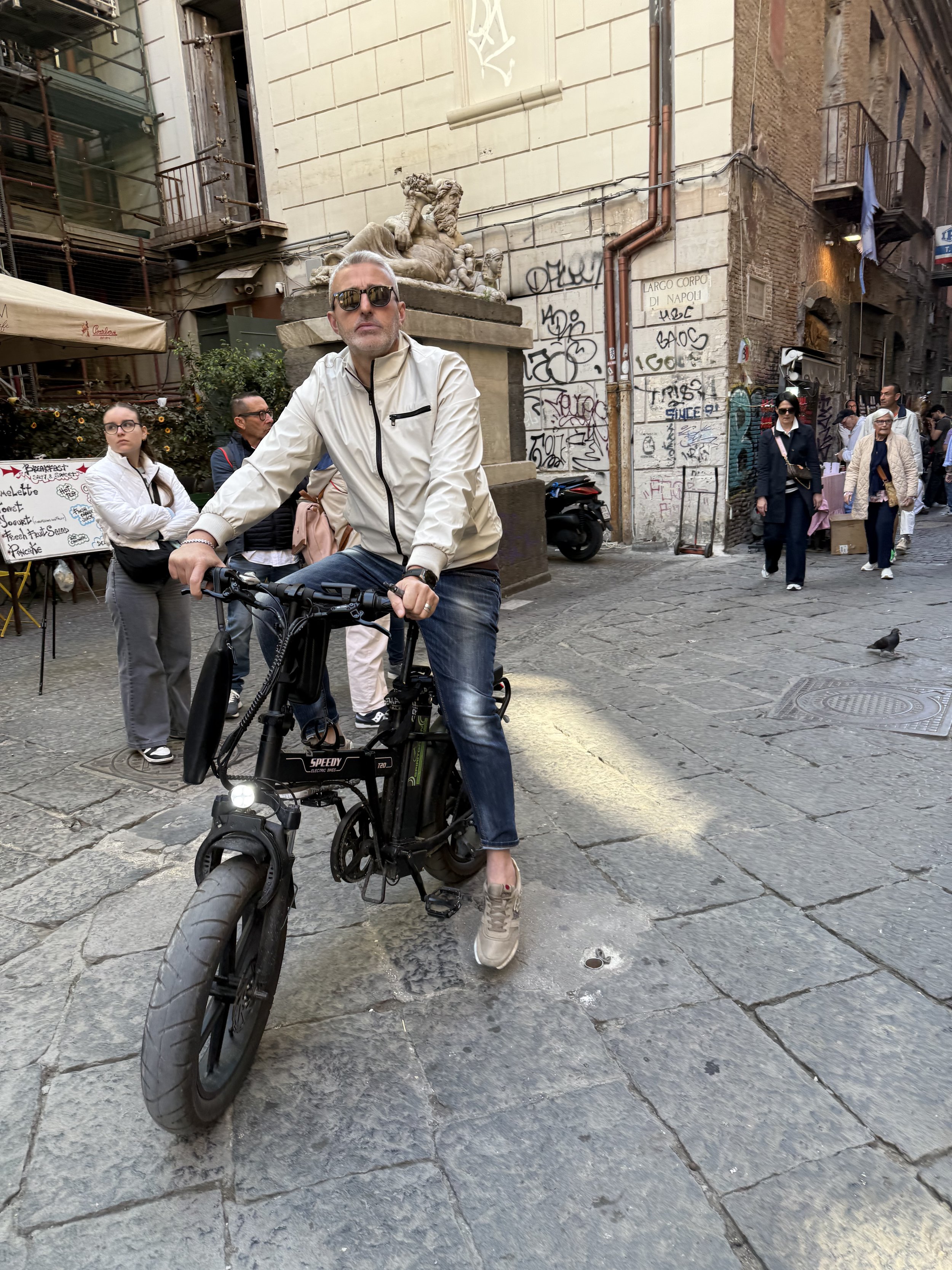 A man with sunglasses and a white jacket on a black electric bike on a cobblestone street in a European city. Behind him, a woman and several pedestrians walk past, with graffiti on the walls and a small lion sculpture above. The scene appears to be 