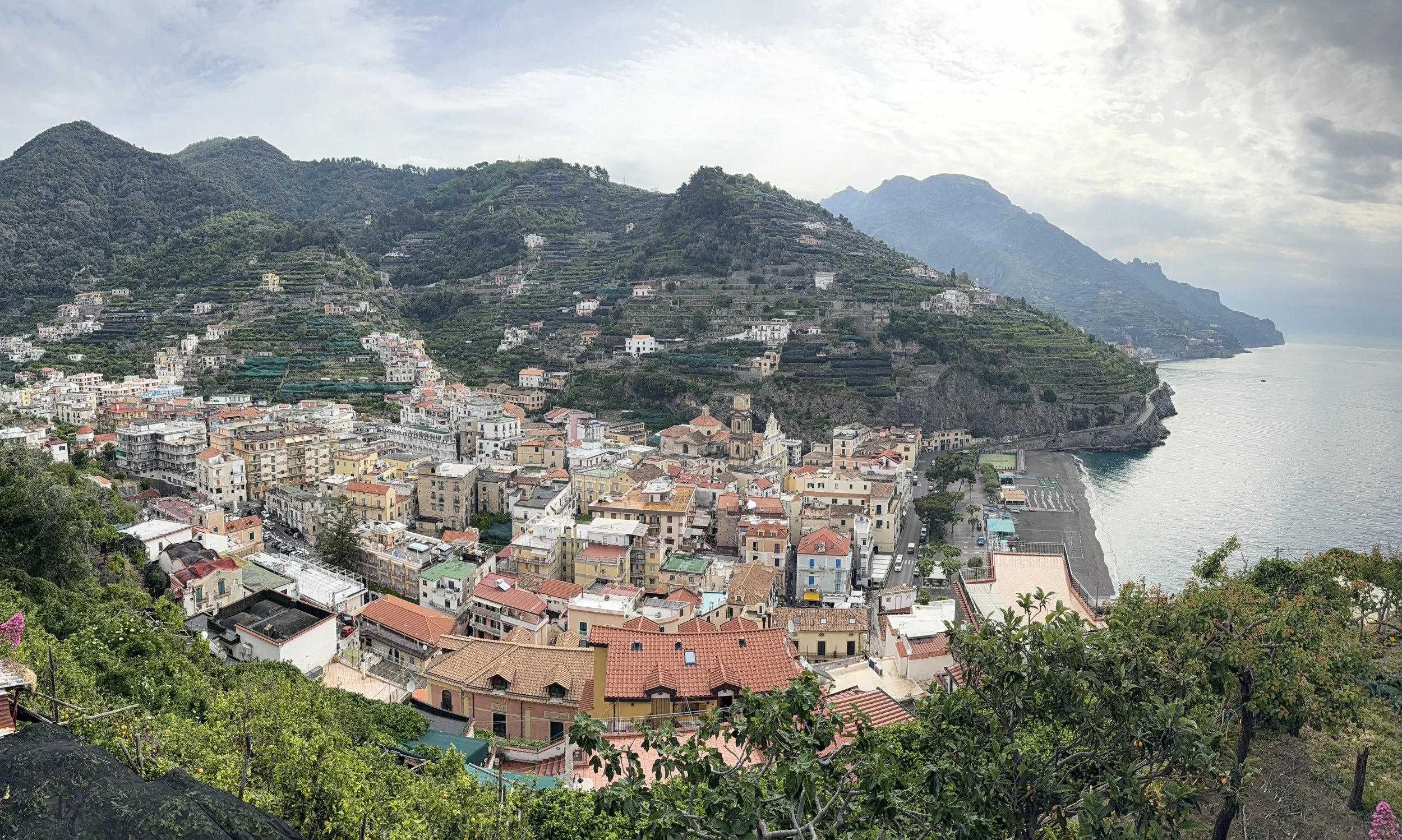 Hilly coastal town with colorful buildings and terraced hillsides overlooking the sea, under partly cloudy sky.