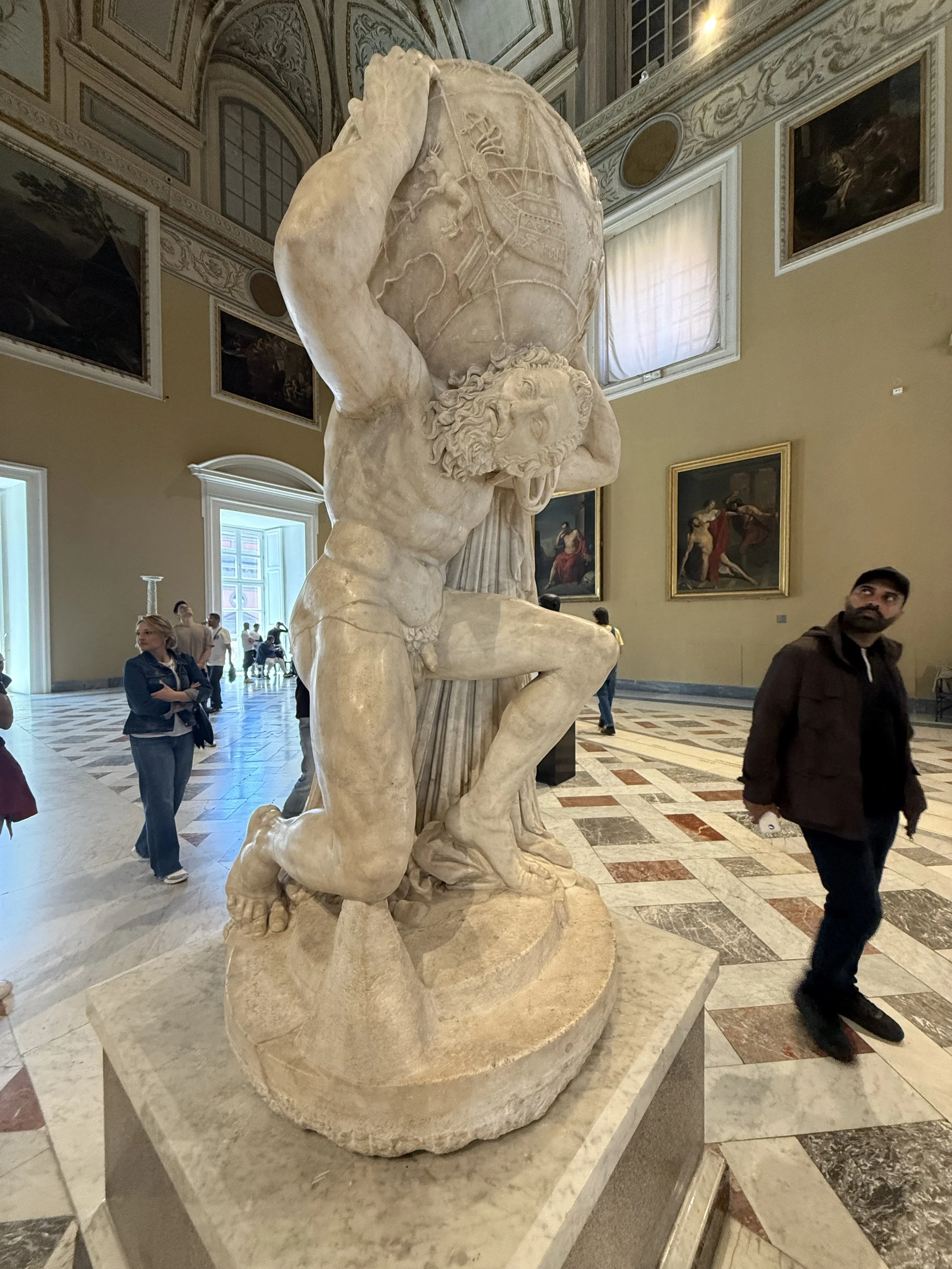 Ancient marble statue of Atlas holding the globe, located indoors among visitors, with paintings on the walls.