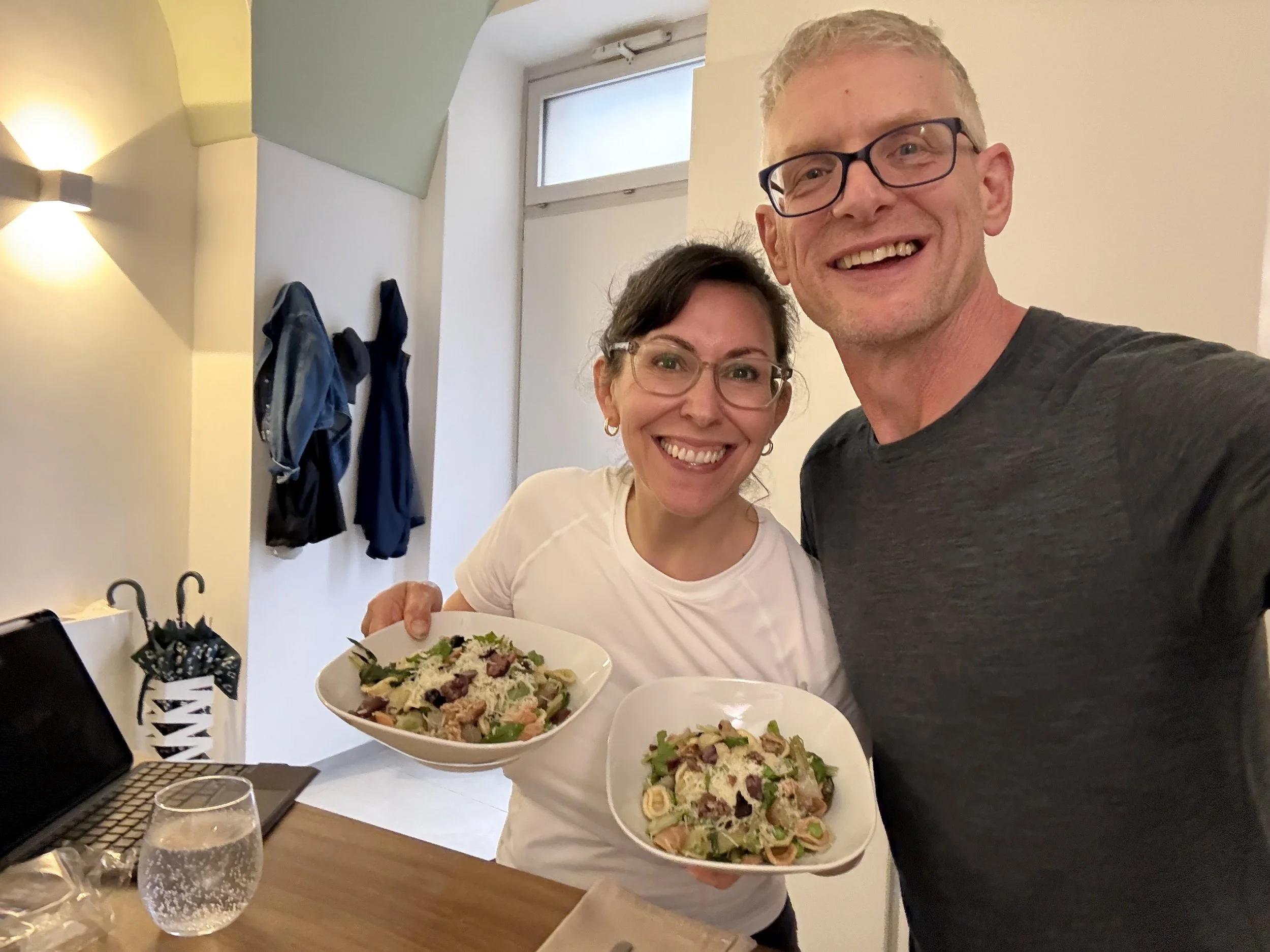 A man and woman smiling, holding plates of pasta salad in a dining area with a wooden table, wine glass, and fluffy drink. The woman wears glasses and a white top, the man wears glasses and a dark shirt, with sunlight coming through a window in the b