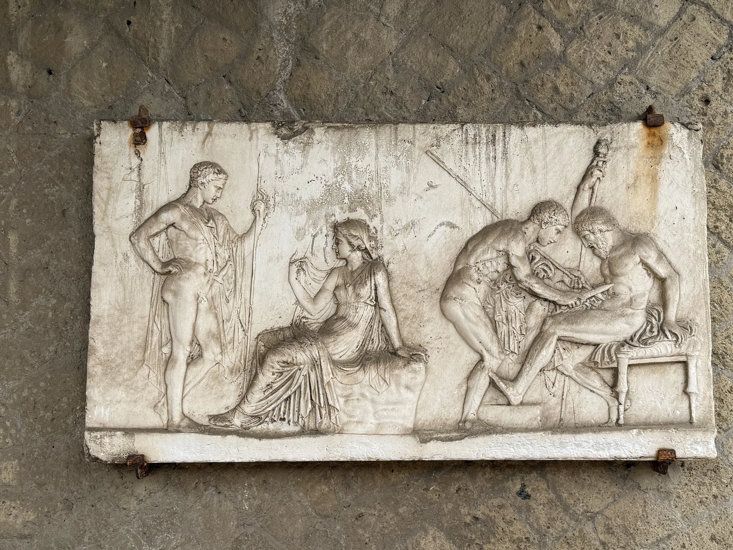 A carved stone relief depicting a scene with multiple ancient figures, including a woman seated and others standing or engaging in various activities, mounted on a stone wall.