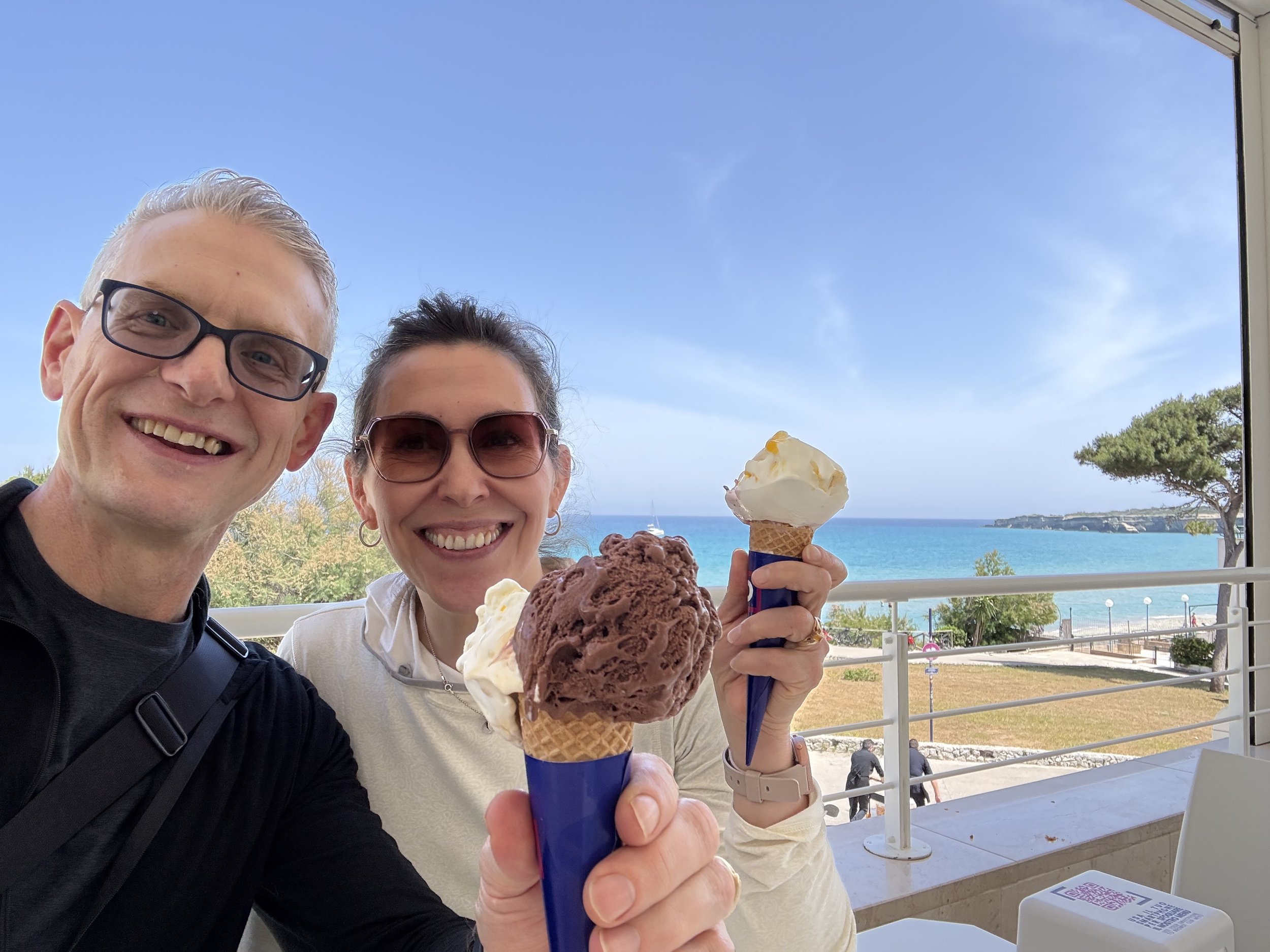 A smiling man with glasses and a woman with sunglasses holding ice cream cones. They are outdoors with a view of the ocean, sky, and trees in the background.