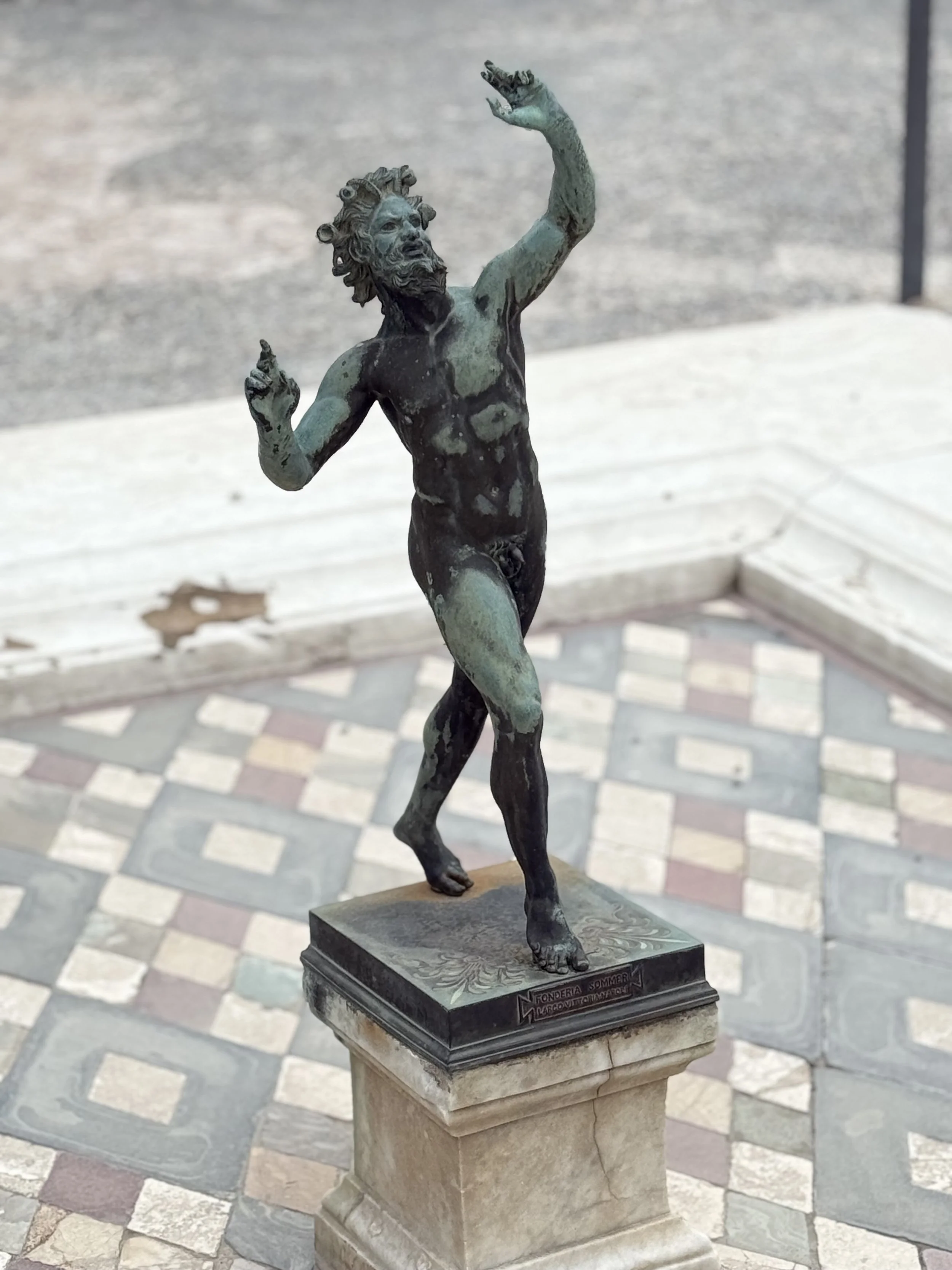 Bronze statue of a man with a beard and long hair, standing with one arm raised and the other bent, on a rectangular base with an inscription. The statue is displayed outdoors on a tiled surface.