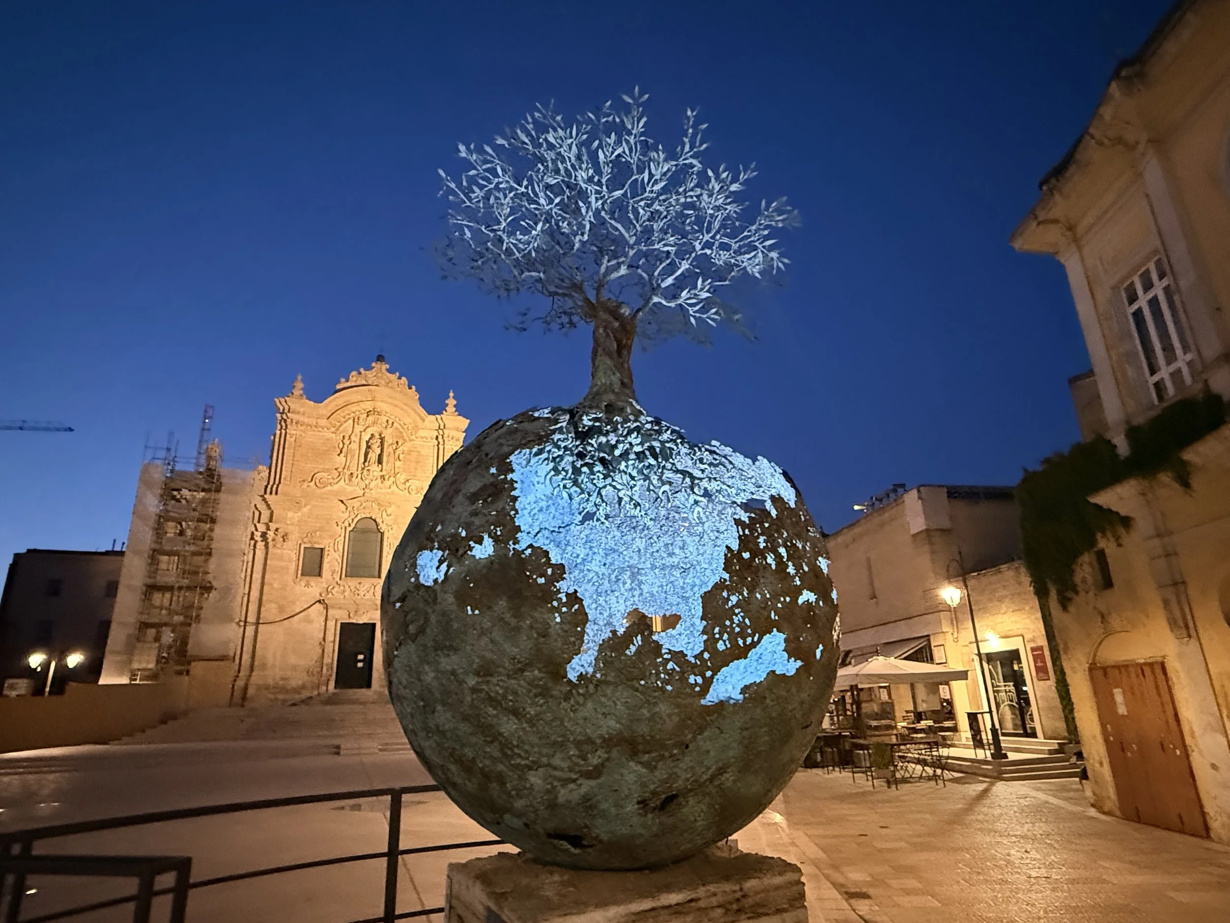 Nighttime scene of a tree with snow or white branches illuminated by blue light in a European-style plaza with historic buildings in the background.
